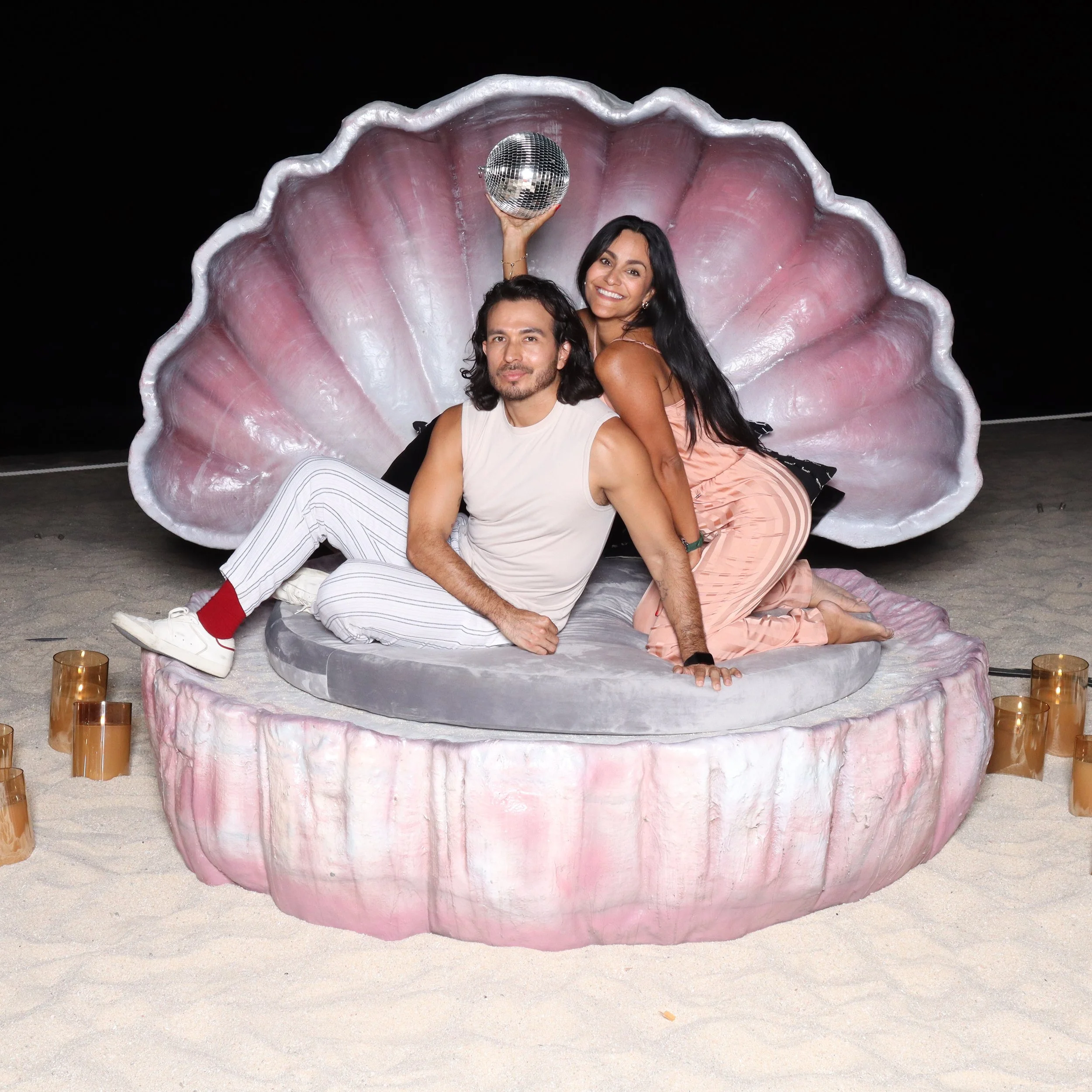 Two people sitting inside a large, pink, shell-shaped seat holding a disco ball, at night with black sky, surrounded by candles.