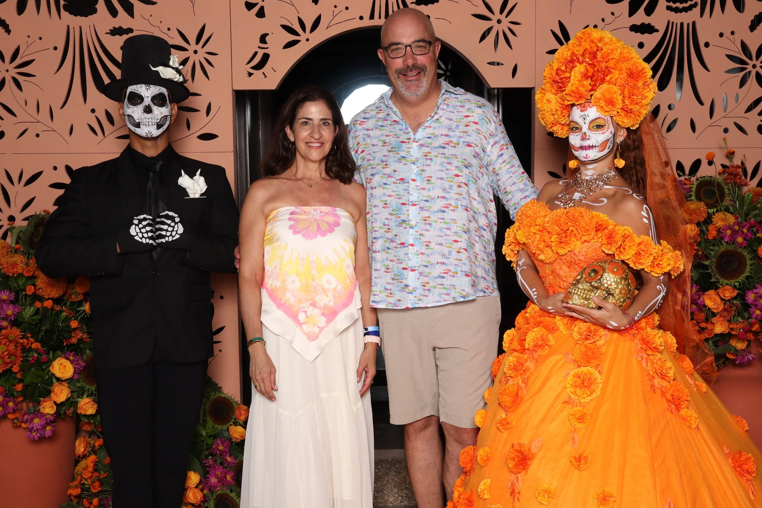 Four people standing in front of a decorative wall, with two individuals dressed in elaborate Day of the Dead costumes, surrounded by vibrant orange and purple flowers.