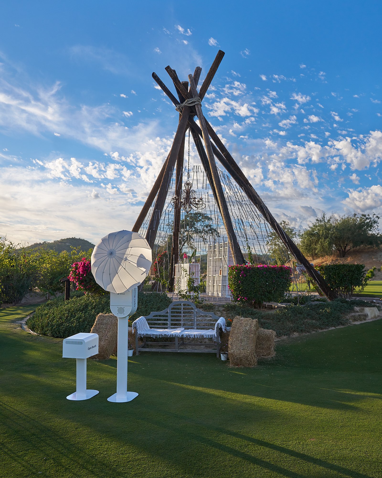 A teepee made of wooden poles tied together with rope, set up outdoors on a grassy area under a blue sky with scattered clouds. There is a white umbrella, a bench with a white cloth, and decorative flowers nearby.