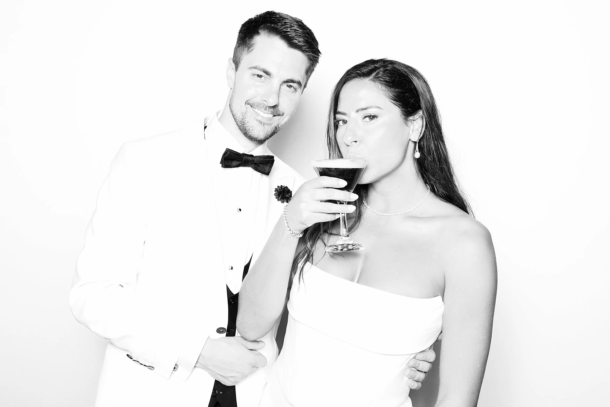 A black-and-white photo of a smiling man in a tuxedo and a woman in a strapless dress, holding a cocktail glass, posing together against a plain white background.