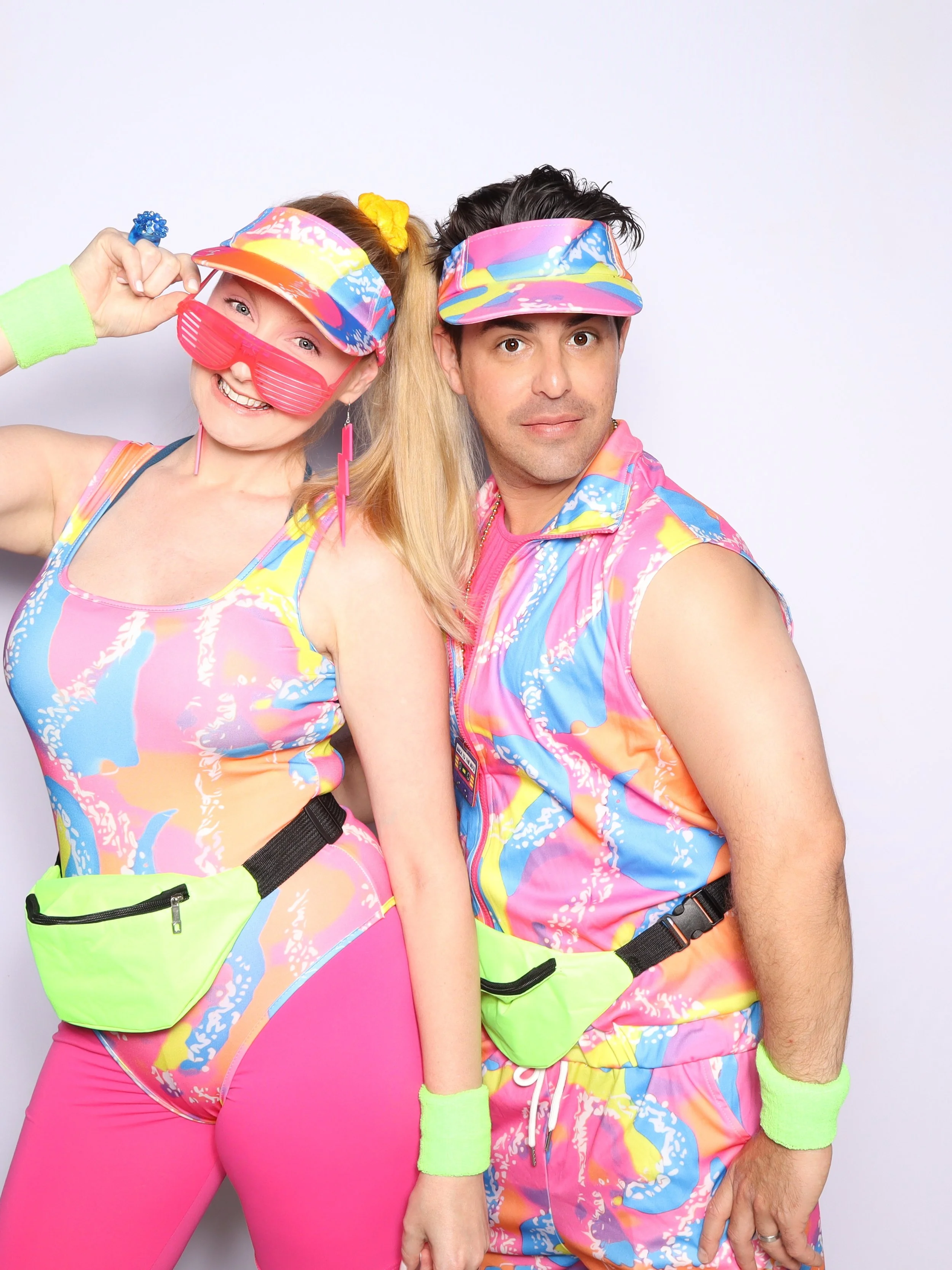 Two people dressed in colorful, retro 80s-style outfits, wearing patterned visors and accessories, standing against a plain white background. The woman is smiling, holding her visor, while the man looks directly at the camera.