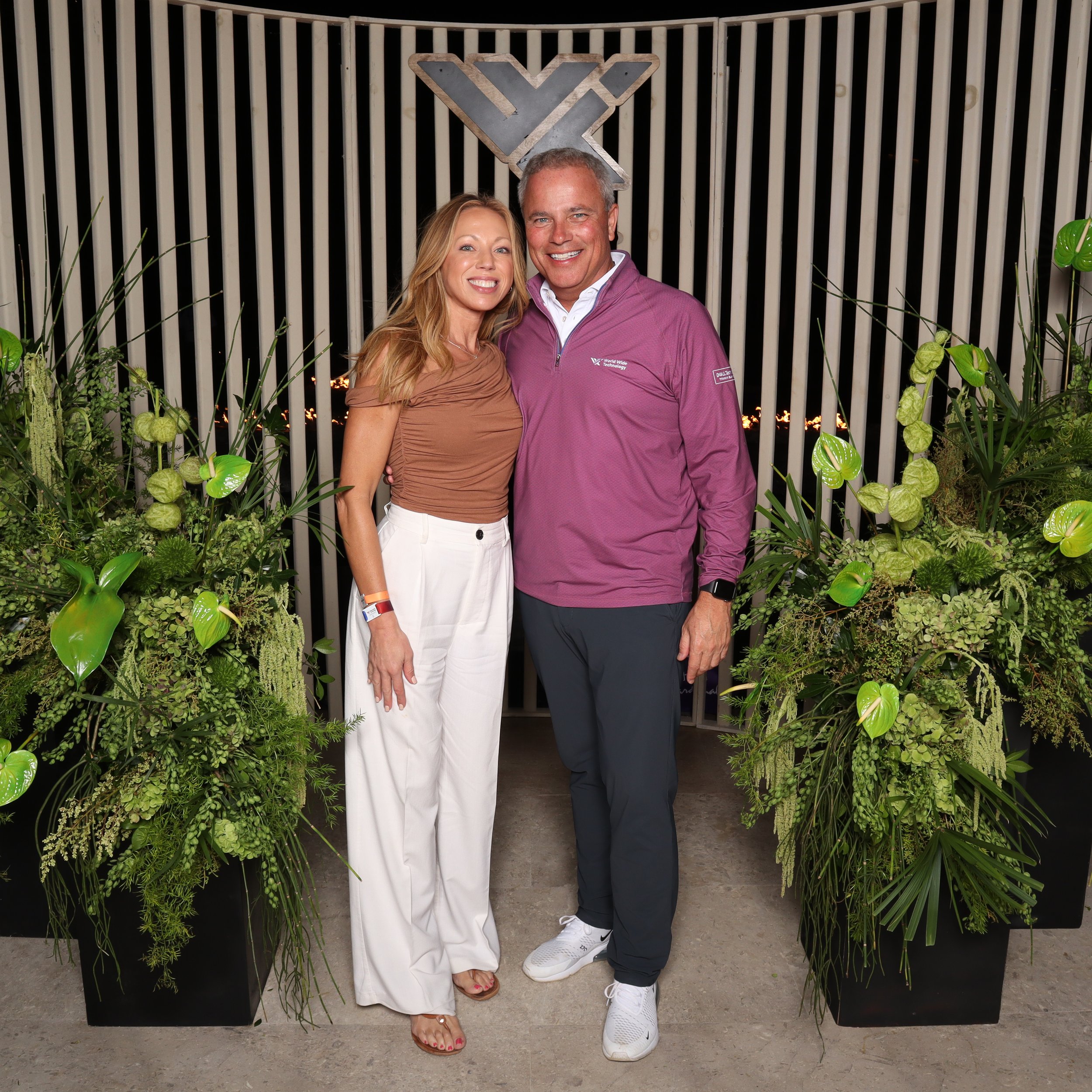 A smiling woman in white pants and a beige top, and a smiling man in dark pants and a maroon shirt, standing together behind a lush green plant arrangement. They are in front of a black metal backdrop with a logo that looks like a stylized 'Y' or 'X'