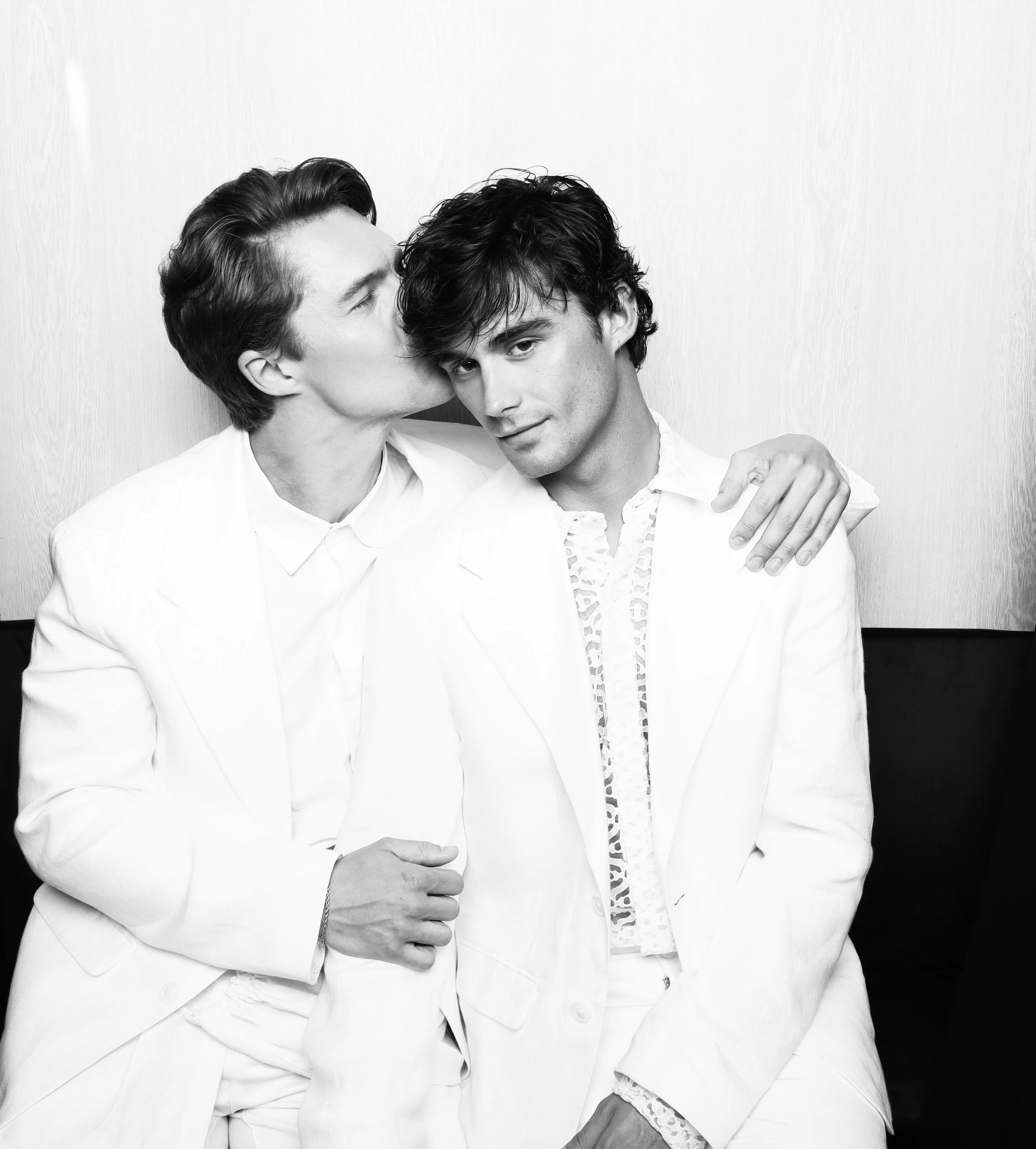 Two men in white suits hugging and sharing a kiss on the cheek, black and white photograph with plain background.