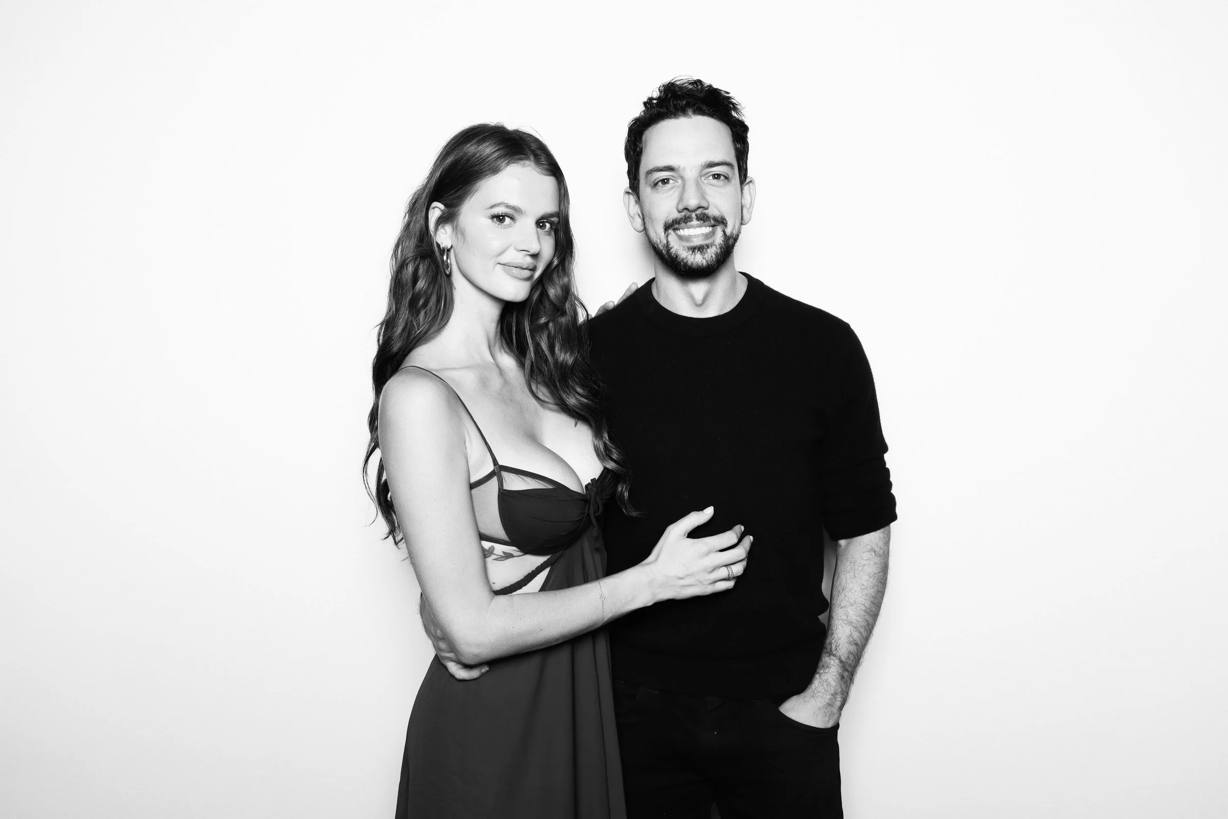 Black and white photo of a smiling woman with long wavy hair wearing hoop earrings and a spaghetti strap top, standing next to a man with short dark hair and a beard, wearing a black long-sleeve shirt, against a plain white background.