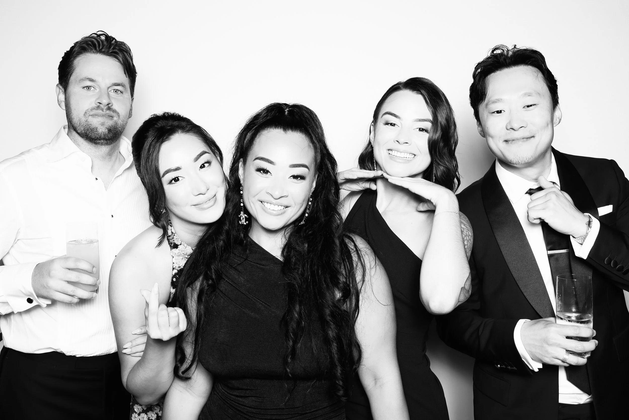 Group of five people dressed in formal attire at a social event, smiling, with some holding drinks, posed against a plain white background.