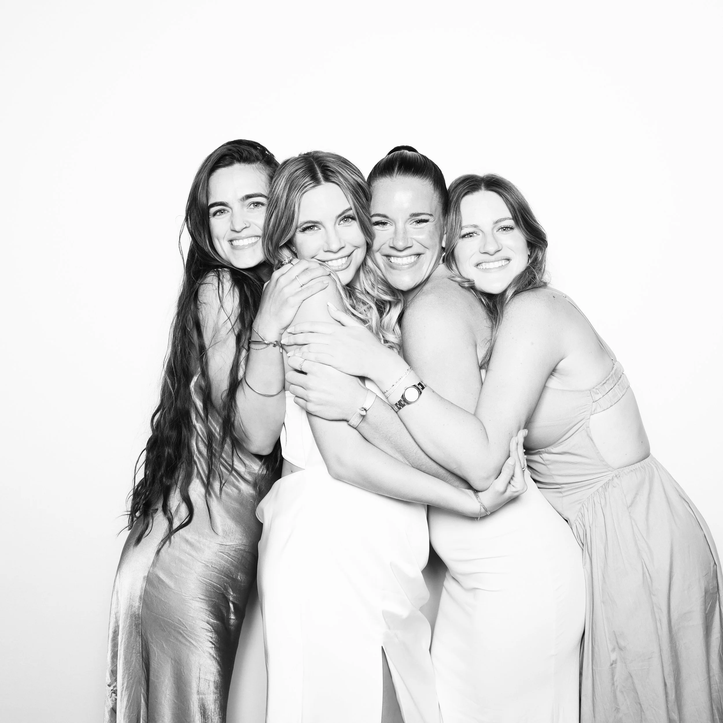 Black and white photo of four smiling women hugging each other against a plain white background.