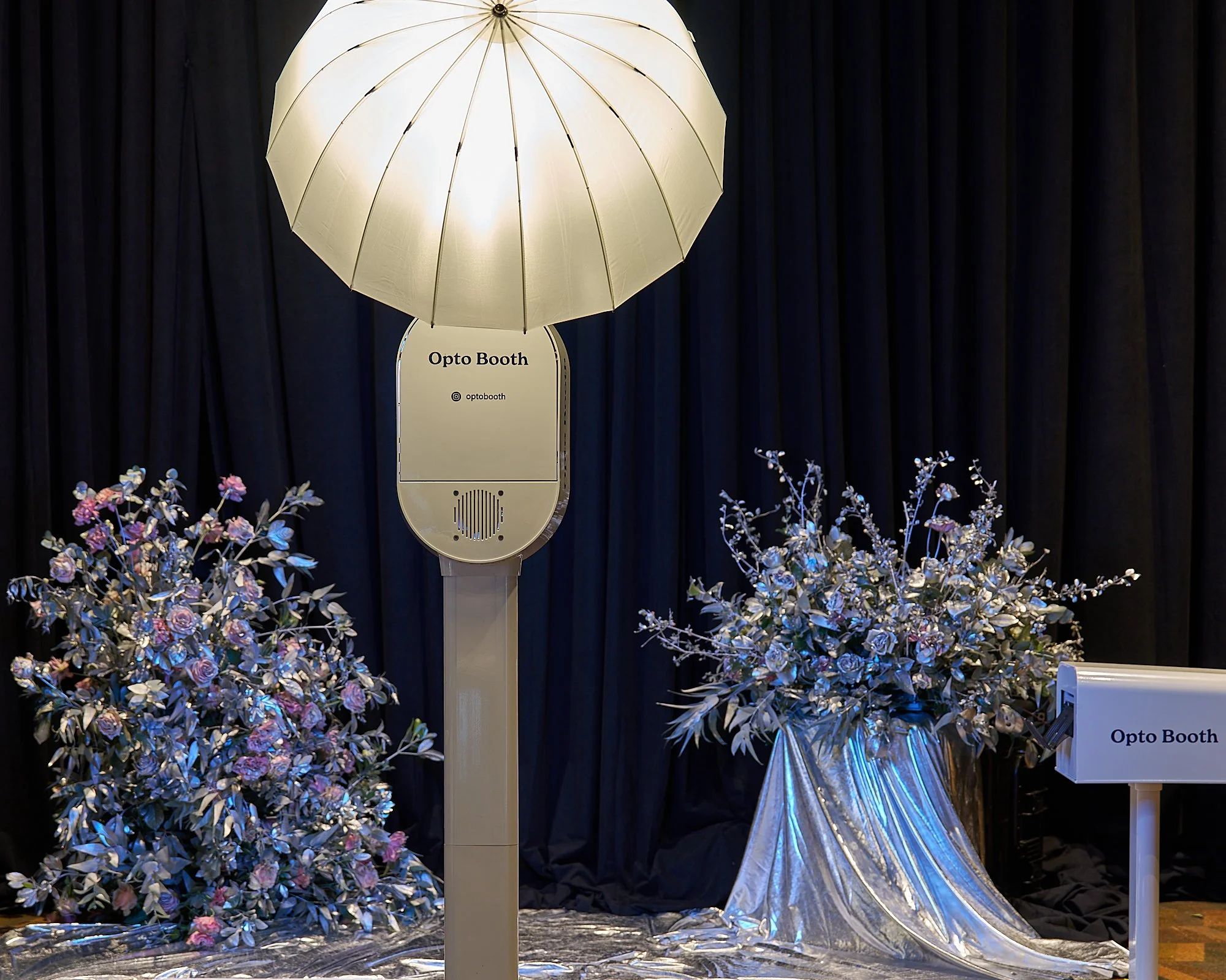 A large, lamp-shaped structure labeled "Opto Booth" with surrounding floral arrangements and dark curtains in the background.