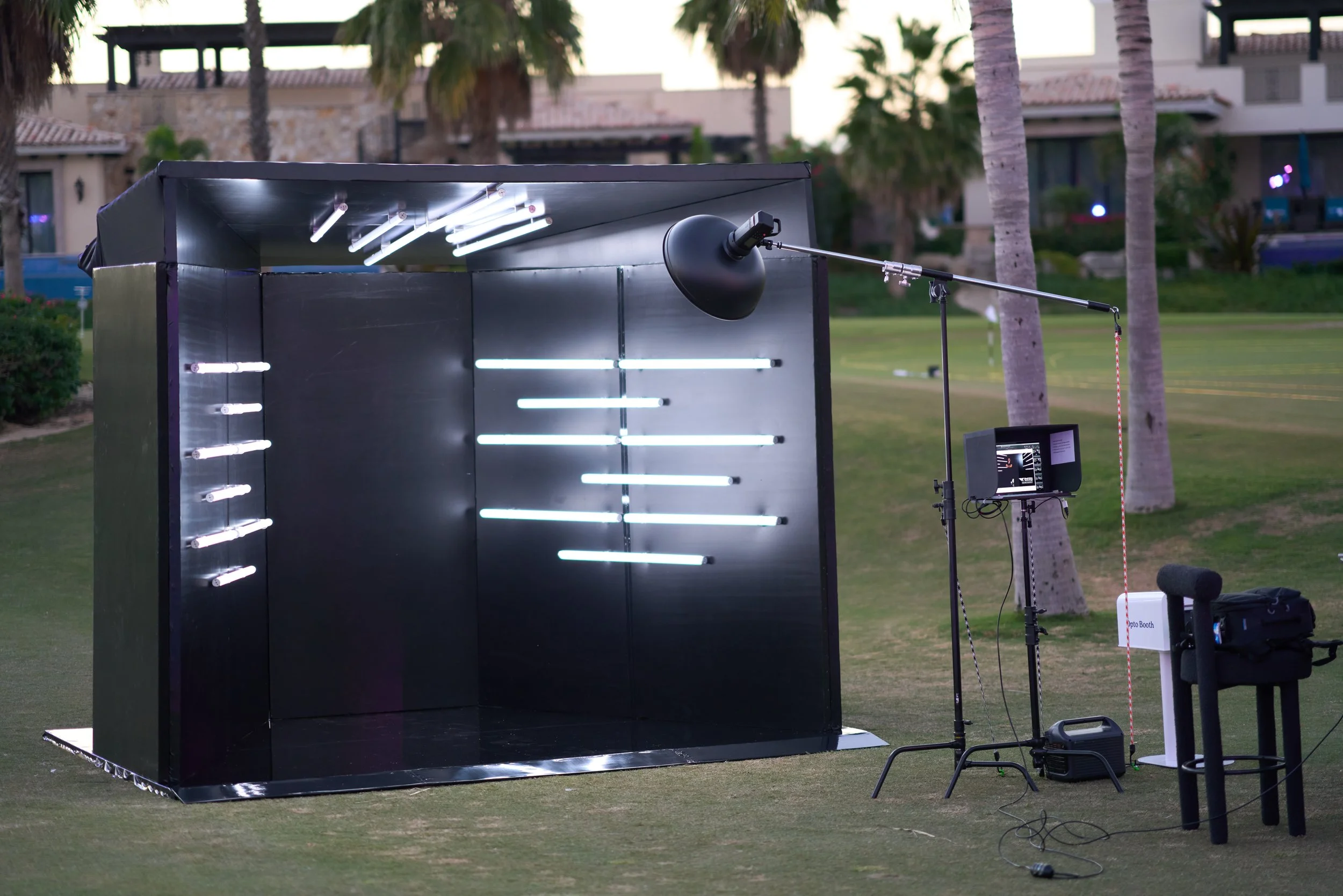 A photo booth setup outdoors on grass, with a black interior and white fluorescent lights, surrounded by palm trees and residential buildings in the background.