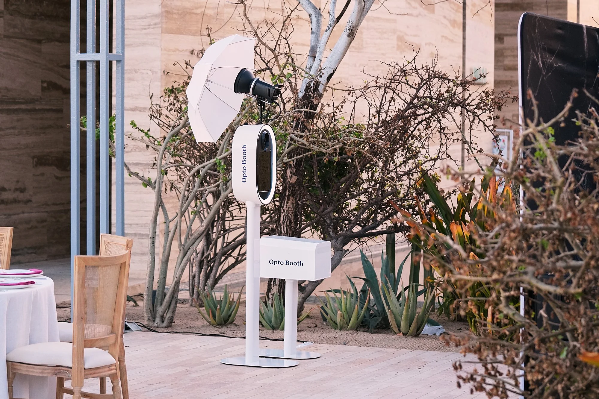 Photo of an outdoor Opto Booth setup, including a white stand with a digital screen, a white umbrella, and a camera mounted on a tripod, surrounded by plants and trees, with a wooden wall in the background.