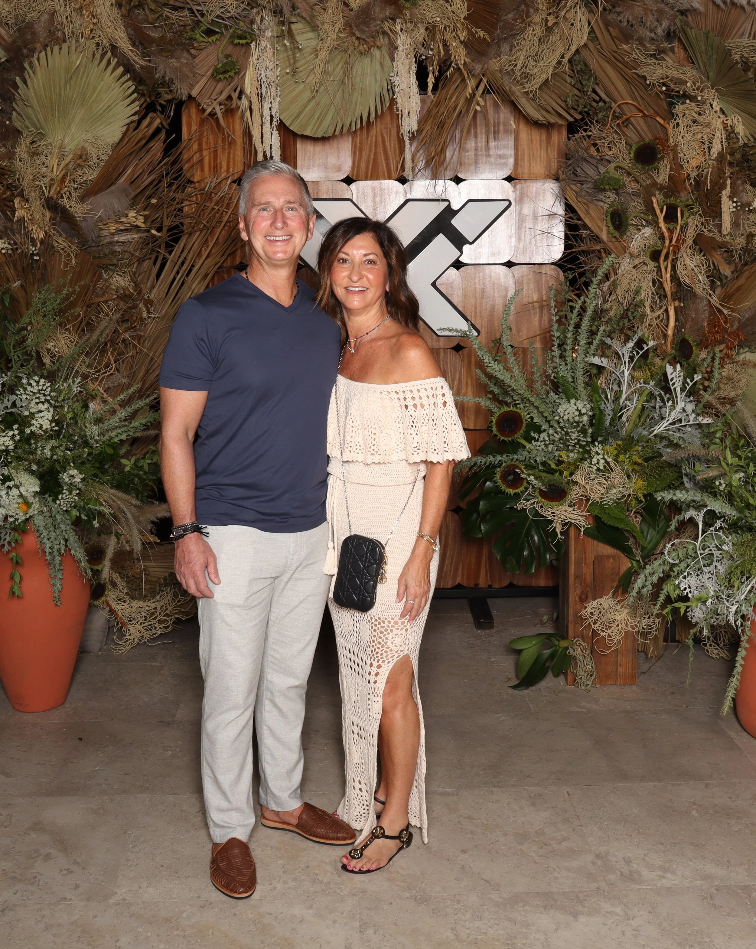 A smiling man and woman standing together in front of a botanical backdrop with large plants and dried foliage, posing for a photo at an event.