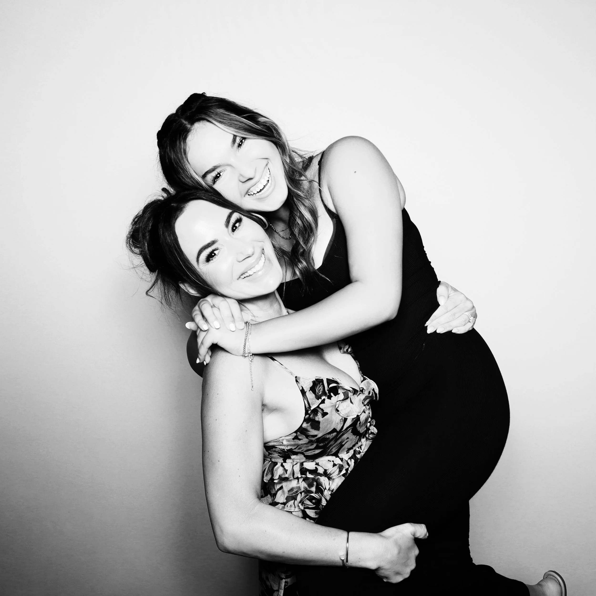 Black and white photo of two women smiling and hugging, one woman with long dark hair wearing a floral dress, and the other with wavy hair wearing a black tank top.