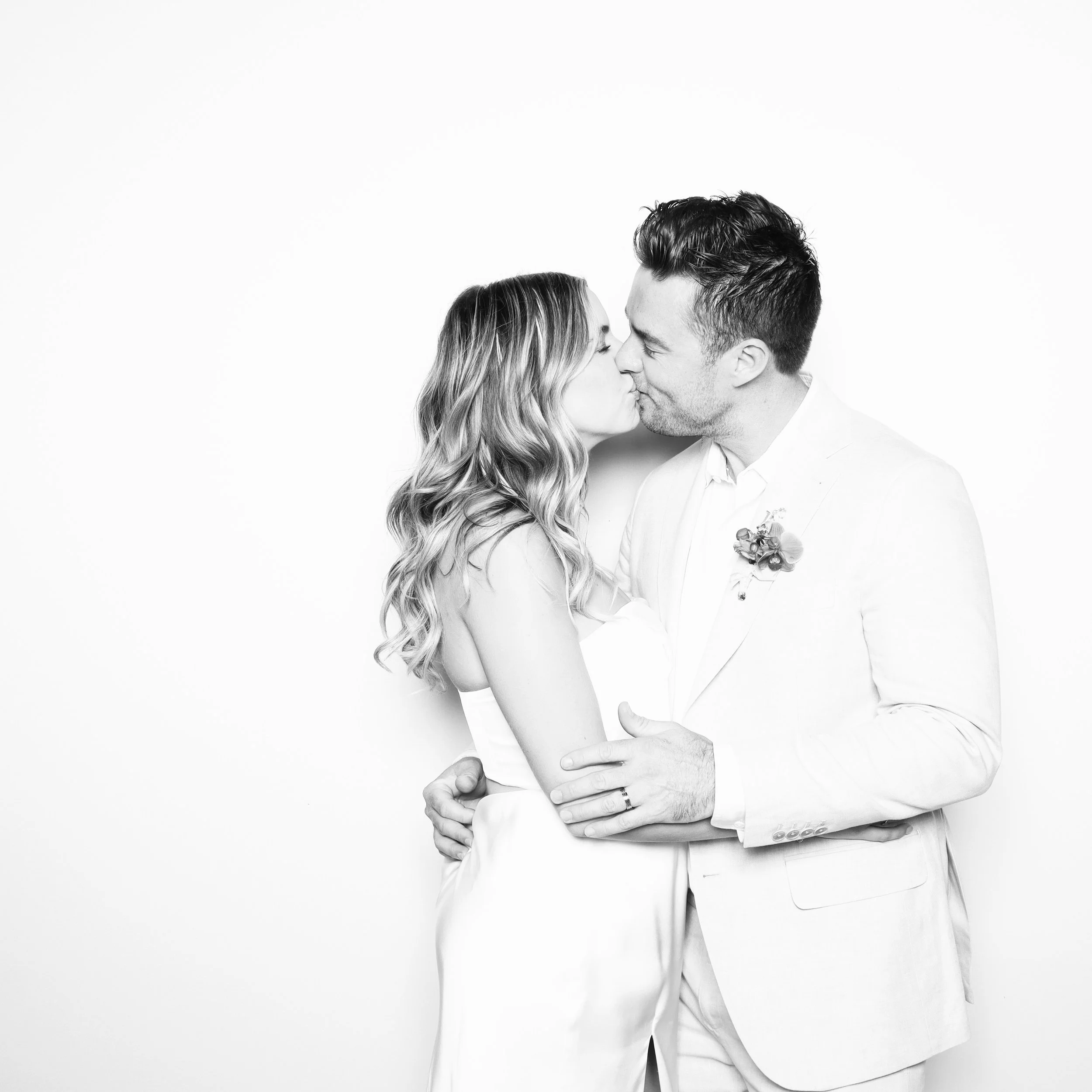 A black and white photo of a couple kissing, both dressed in formal white attire, with the man's arm around the woman's waist.