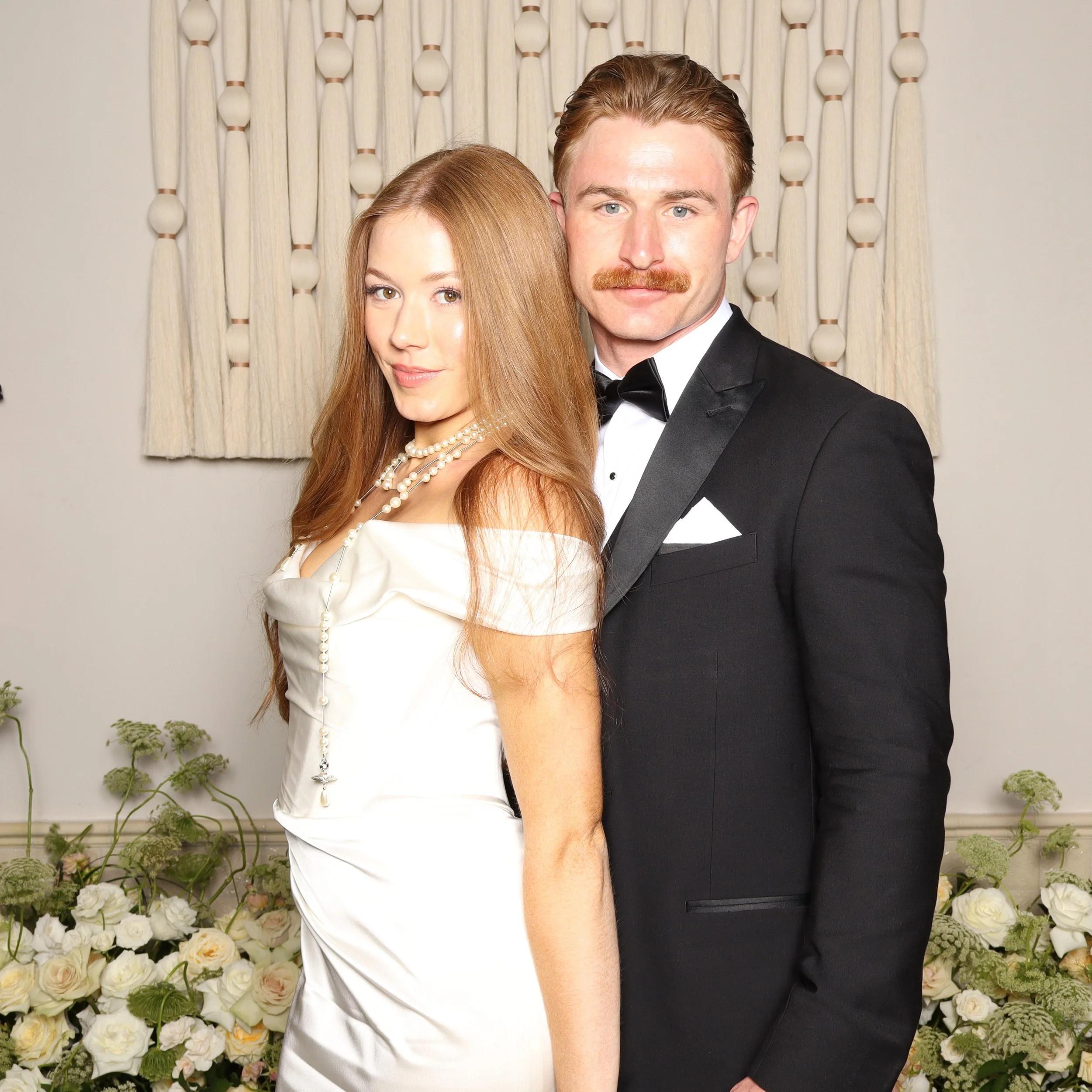 A man and a woman dressed in formal attire pose in front of a wall decorated with white and peach-colored flowers and a curtain. The woman has long red hair and wears a white dress with pearl jewelry. The man has brown hair, a mustache, and is wearin