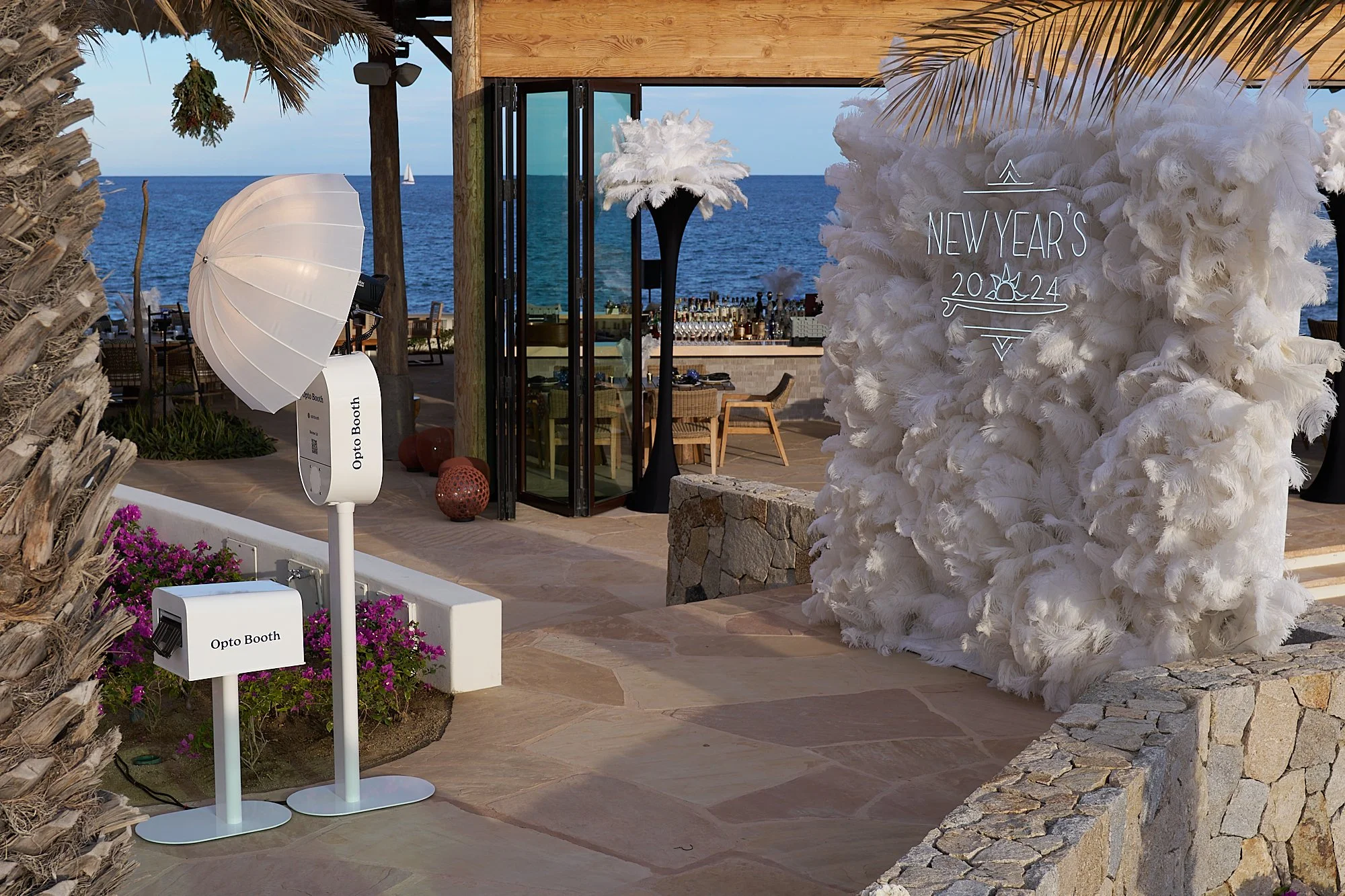 A seaside bar area decorated with large white feather floral arrangements and illuminated New Year's sign for 2024. The area features wood accents, outdoor seating, and ocean views, with a sailboat on the horizon.