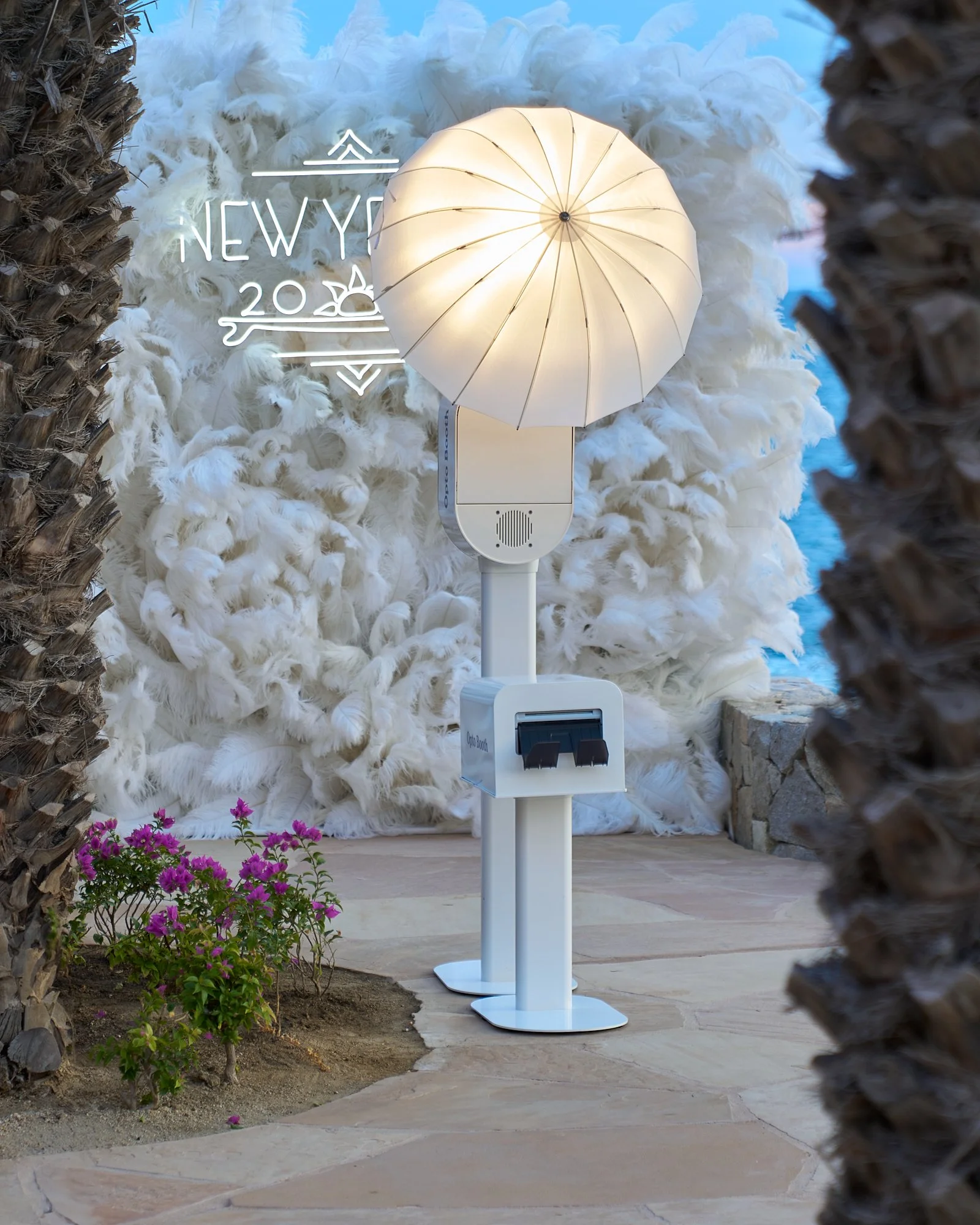 A photo taken in an outdoor area showing a white electric vehicle charging station and a white feathered feather boa backdrop. The scene also features two palm trees on each side and pink flowering plants in the foreground.