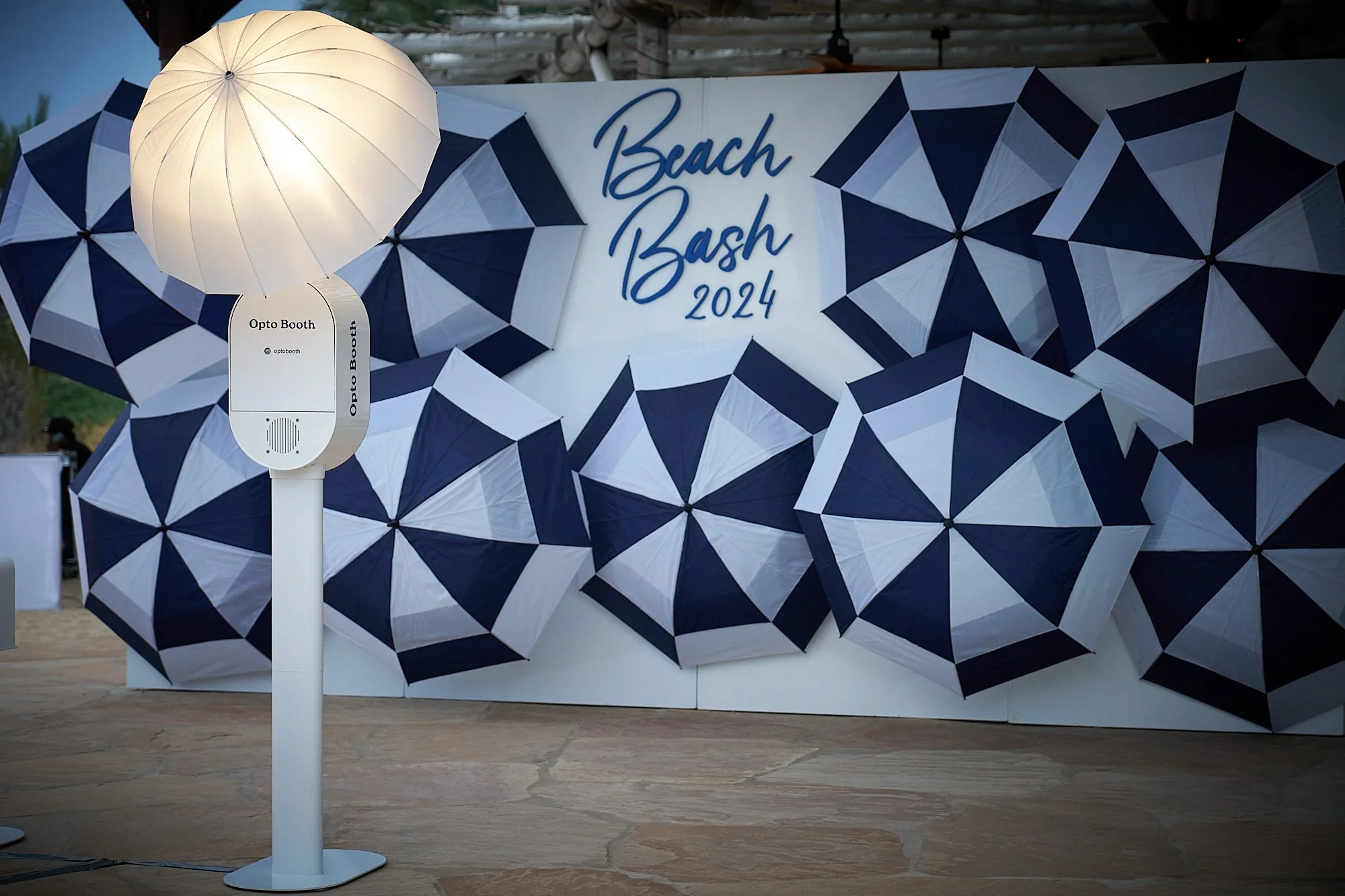 Outdoor display for Beach Bash 2024 with many umbrellas in navy and white, a white illuminated umbrella, and a sign for Opto Booth.