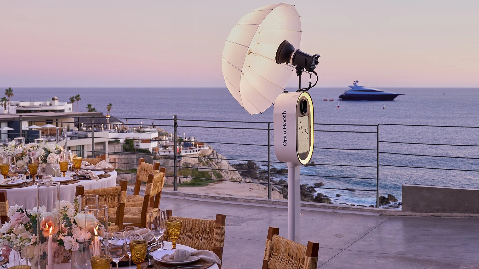 Outdoor event setup with dining tables, floral centerpieces, and glassware near a railing overlooking the ocean, with a yacht in the distance and an Opto Booth photo booth with umbrella lighting.
