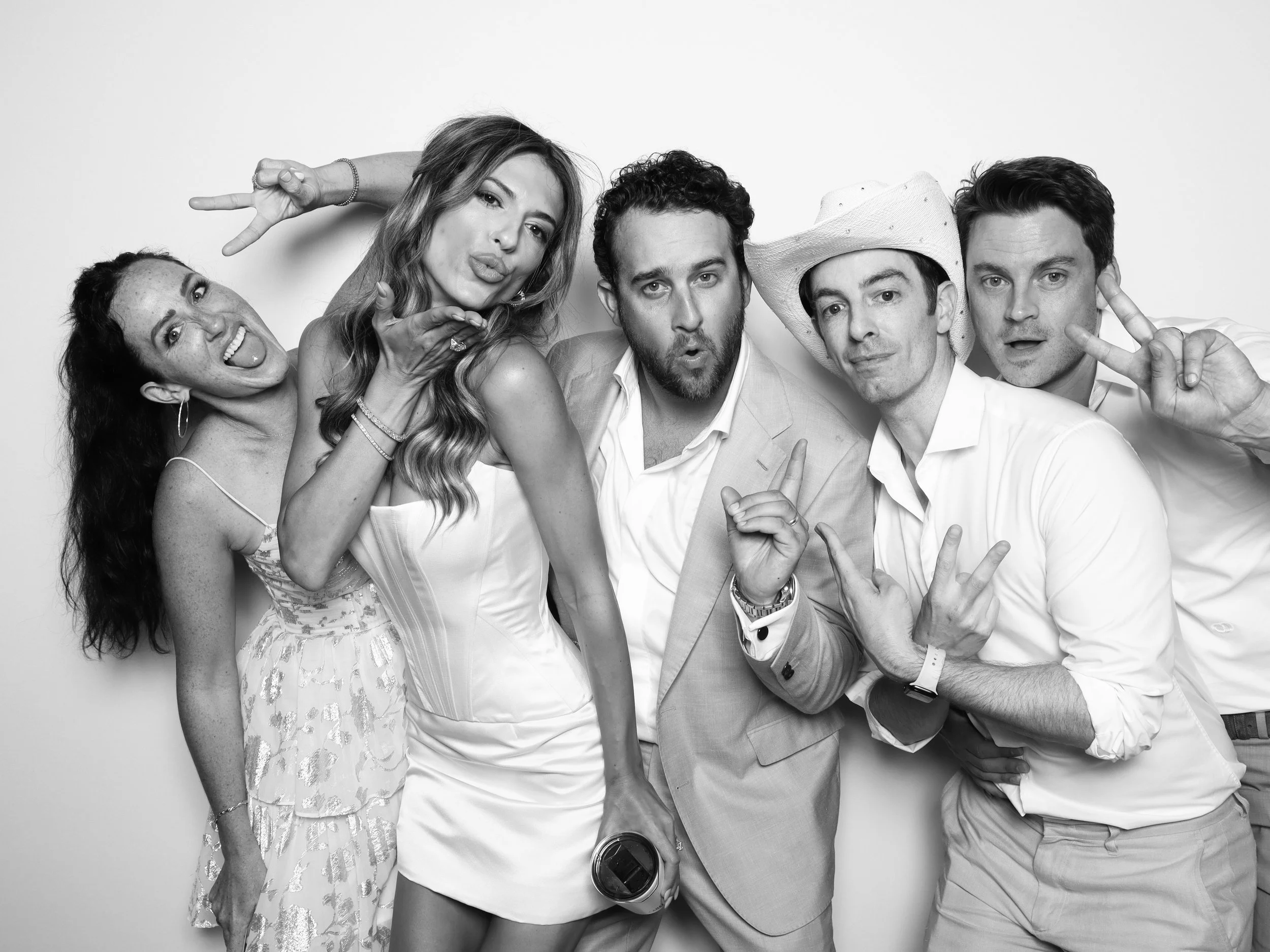 A group of five friends taking a selfie together in black and white. They are smiling and making playful gestures, standing against a plain white background.