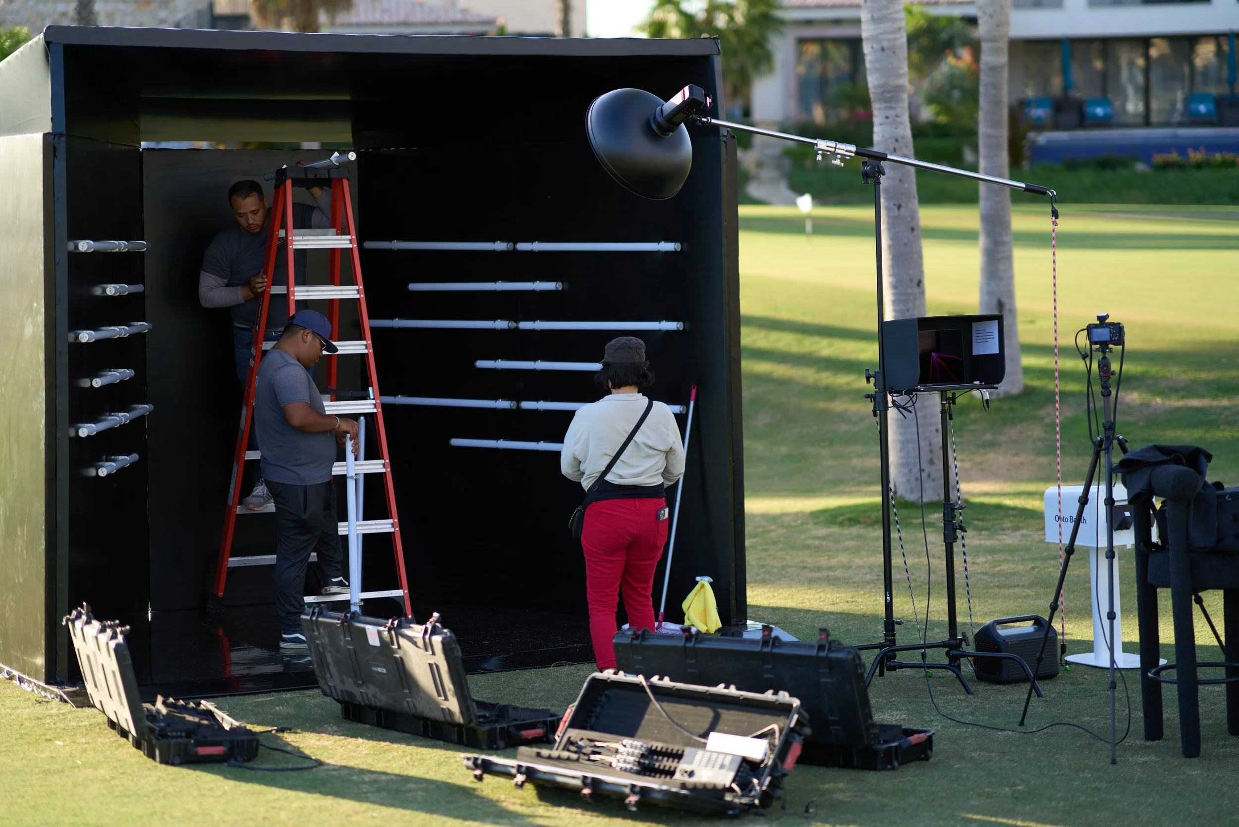A film crew setting up equipment outdoors with a green park and trees in the background. They are working with lighting and camera gear inside a black tent-like structure.