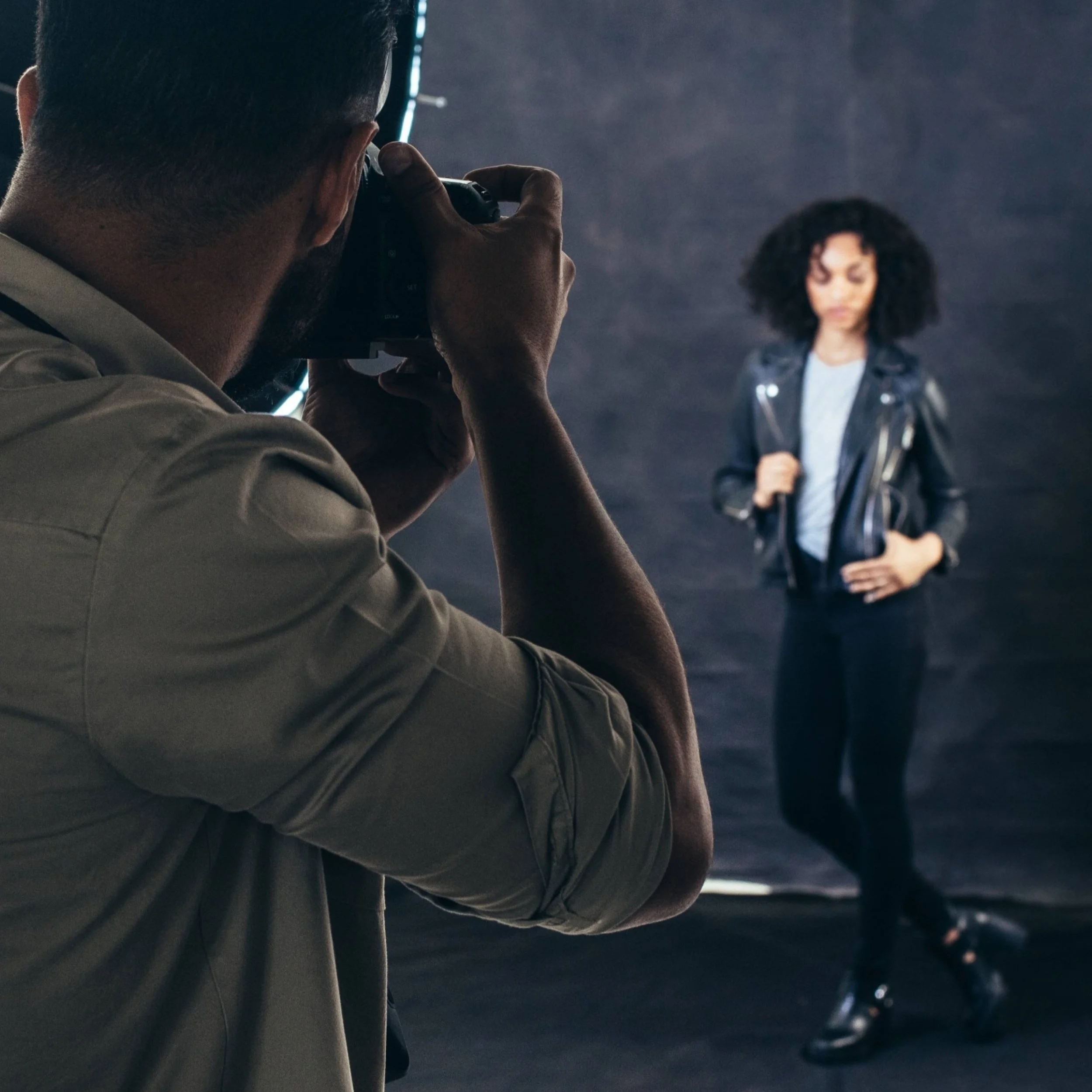 A photographer taking a picture of a woman with curly black hair, standing against a dark background.