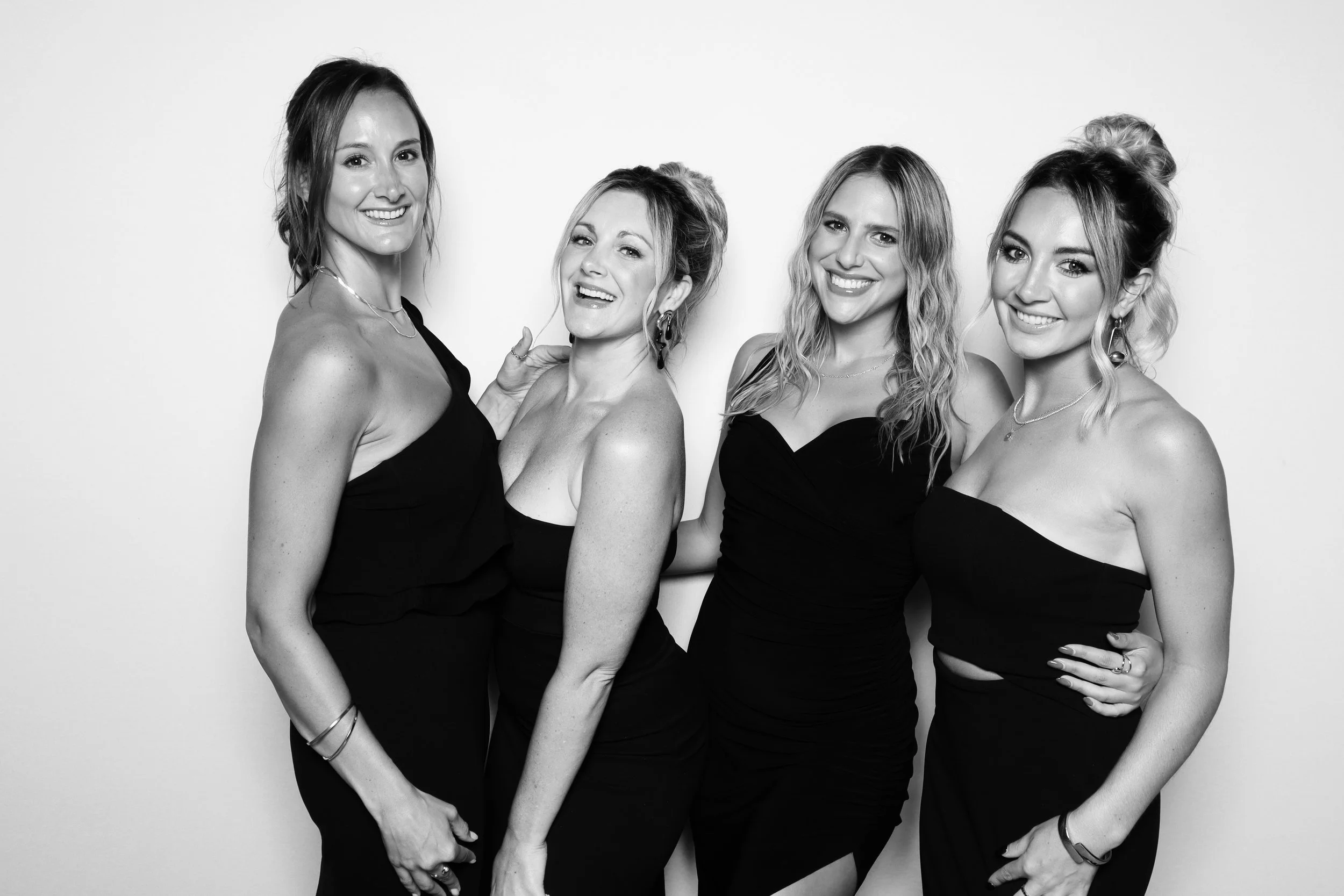 Four women in black dresses smiling and posing together against a plain white background.
