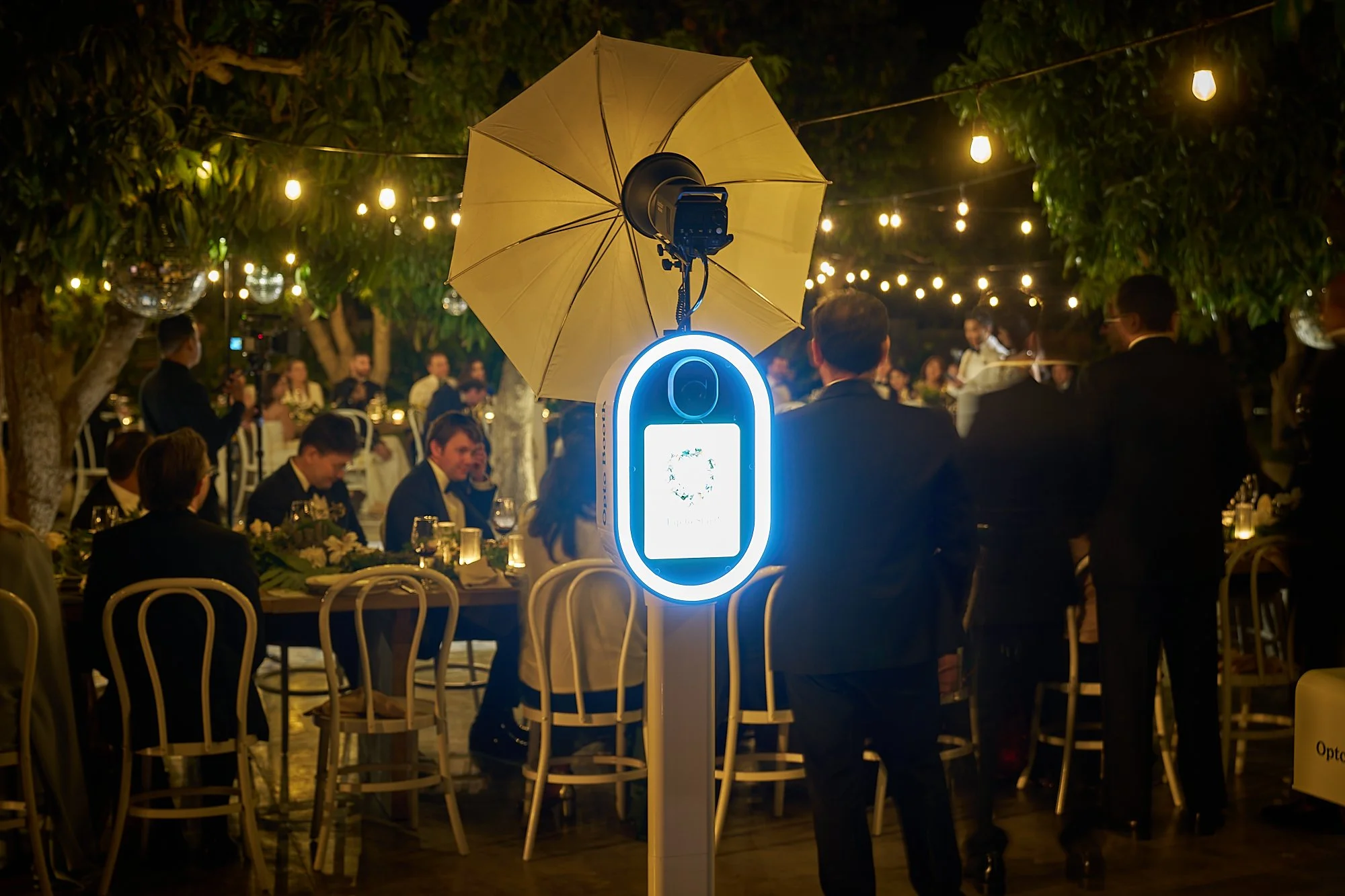 An outdoor evening event with people seated at tables under string lights and trees. A large umbrella is mounted on a stand with a device attached, illuminated by a bright blue light, in front of the gathering.