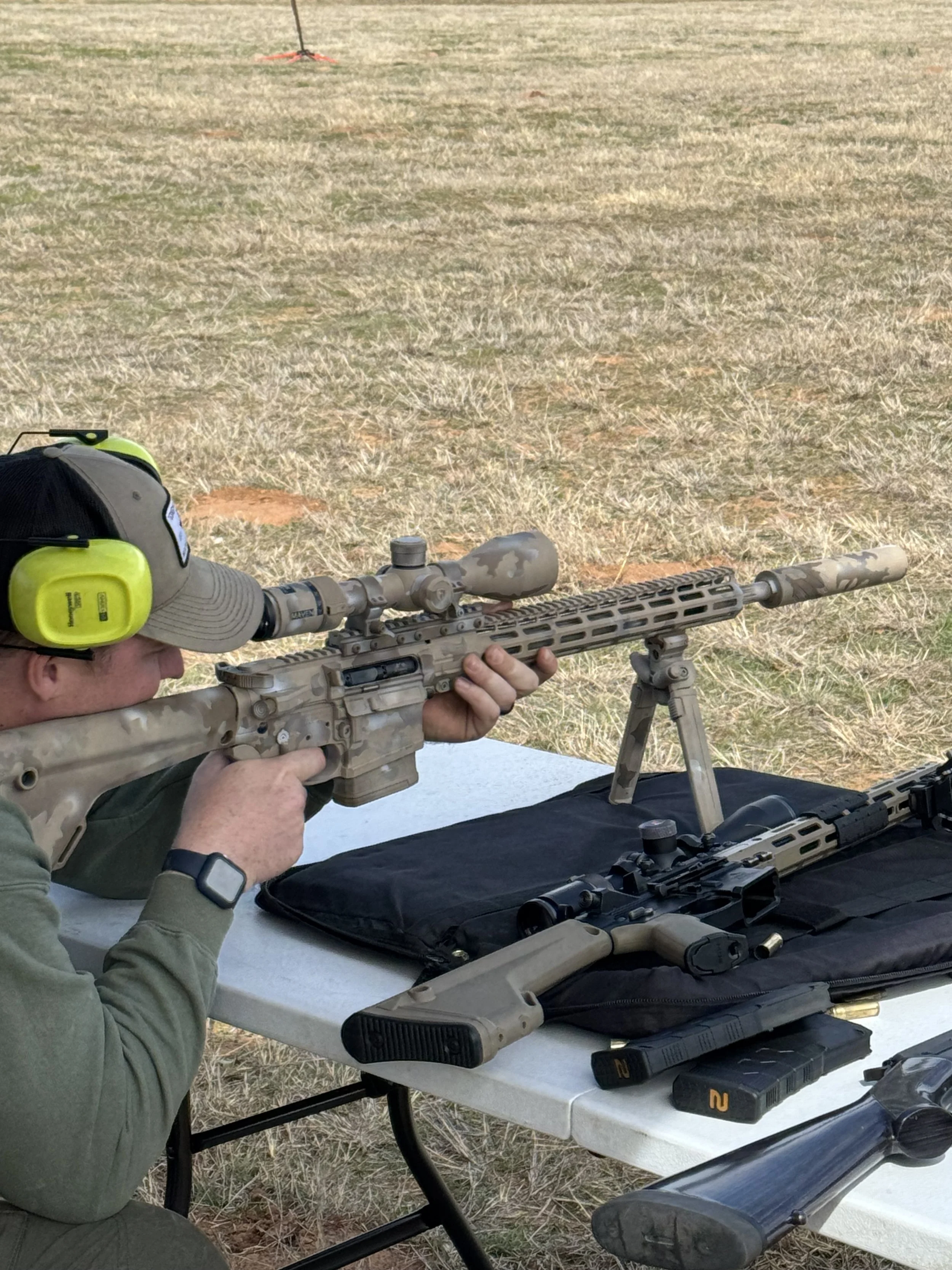 A person aiming a sniper rifle with a scope, lying on a table with additional rifle accessories and magazines, outdoors on dry grass.