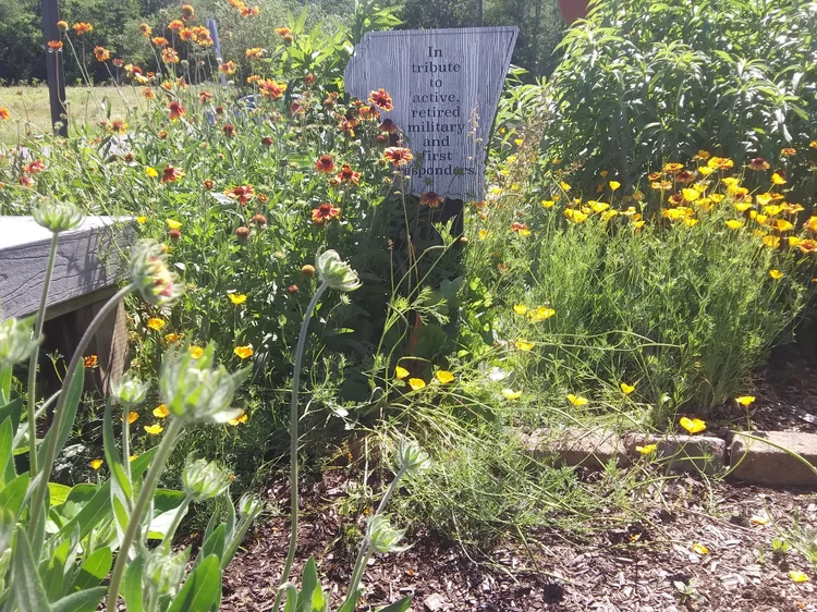 A garden with various yellow and orange flowers, a wooden sign in the background reading 'In tribute to active, retired military and first responders,' and a pathway with bricks on the right side.
