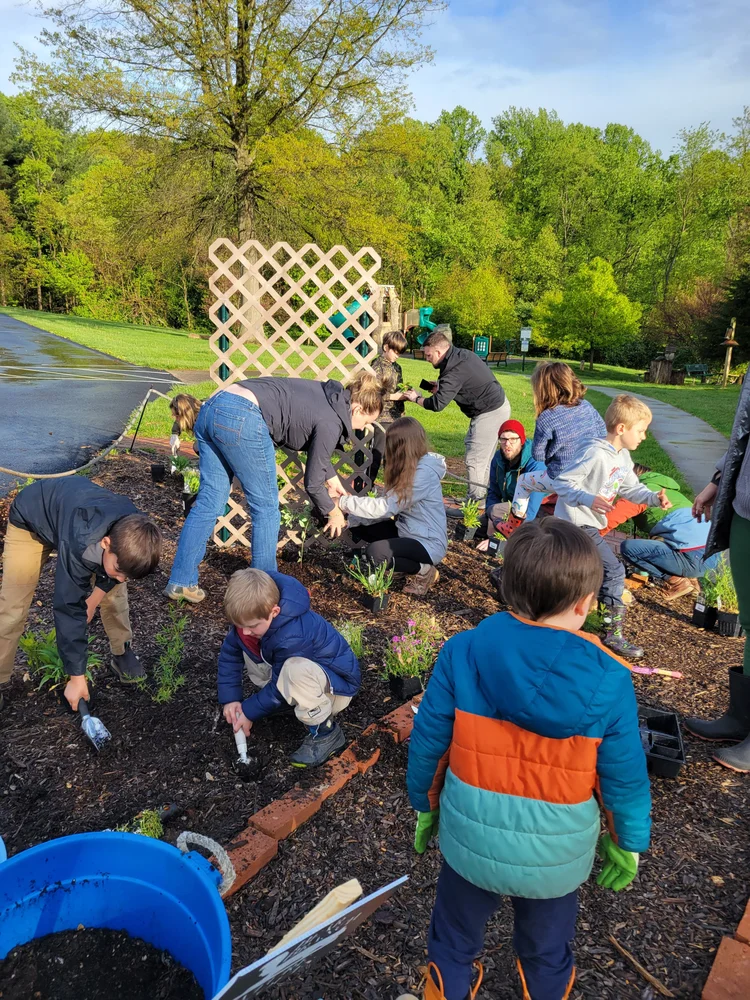 Children and adults planting flowers and shrubs in a garden bed outdoors, with greenery and trees in the background.