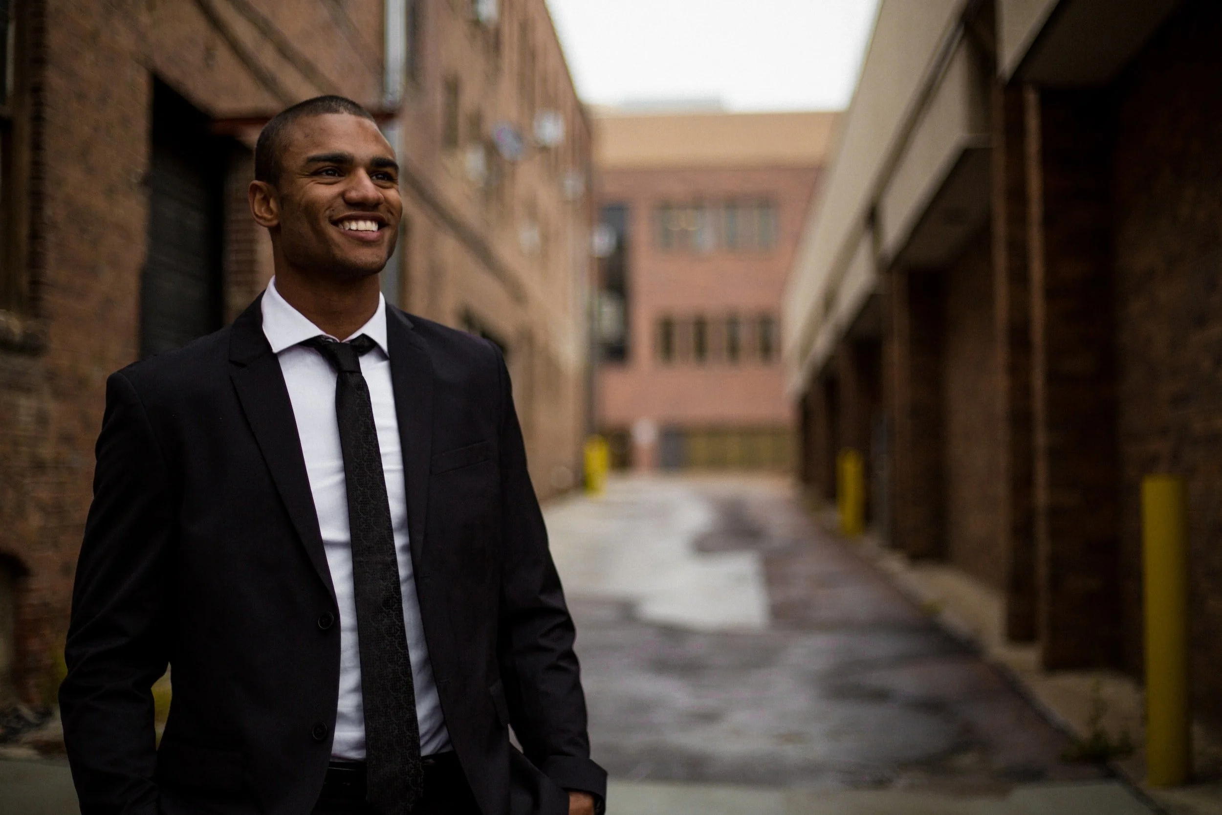 A smiling man in a black suit and tie standing in an alleyway with brick buildings on either side.