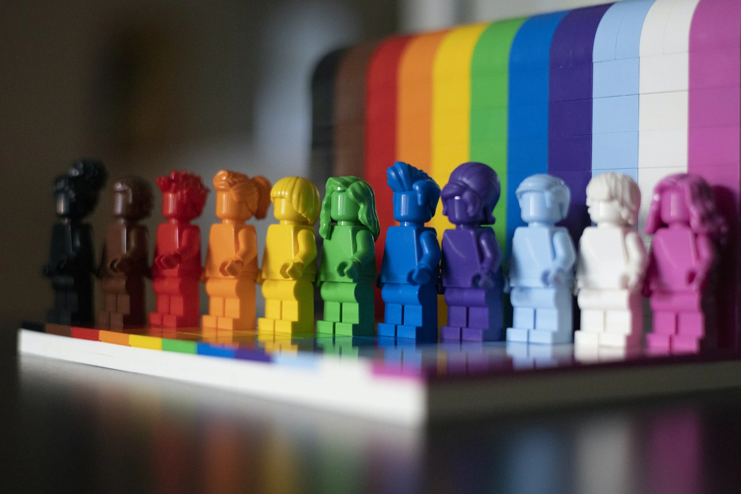 Colorful LEGO minifigures arranged in rainbow order from black to pink, with a rainbow-colored LEGO tile set in the background.