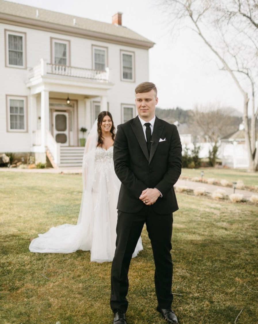 A bride and groom outside a white house, with the bride smiling and the groom looking serious. The bride is wearing a white wedding dress with a veil, and the groom is in a black suit and tie.