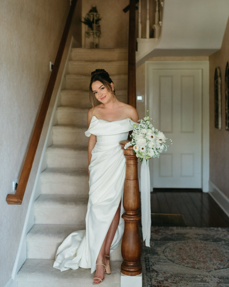 A woman in a white strapless wedding gown standing on a staircase, holding a bouquet of white flowers, in a cozy indoor setting.