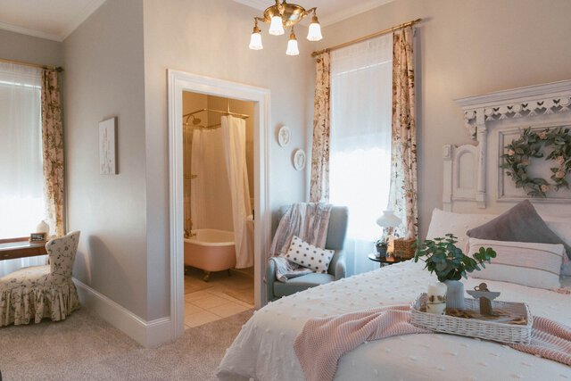 A cozy bedroom with floral curtains, a white bed with decorative pillows, and a tray with plants and a candle. There's a sitting area with an upholstered armchair, a small table, and a window with sheer curtains. An en-suite bathroom with a bathtub and shower is visible through an open door.
