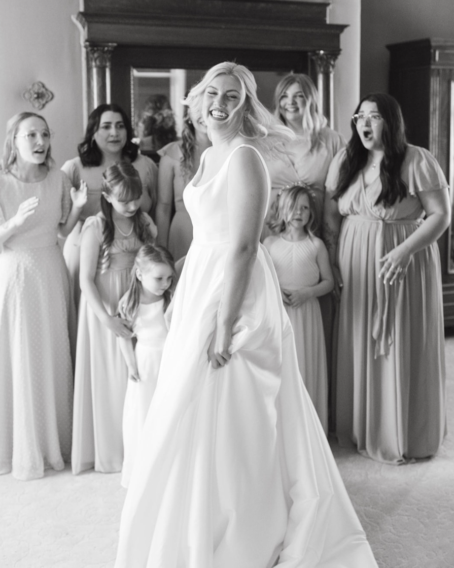 A bride in a wedding dress smiling and holding the skirt of her dress, surrounded by a group of women and girls in formal attire, inside a room with wooden furniture.