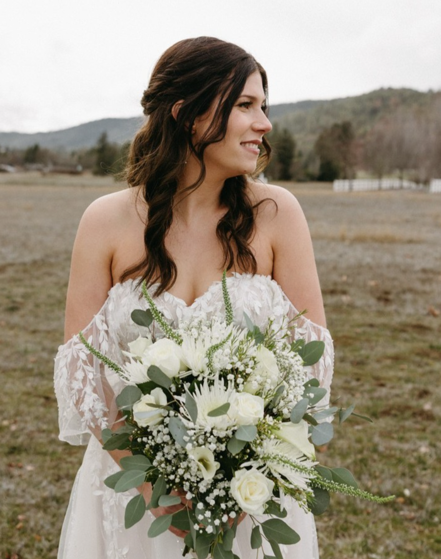 Bride in a white wedding dress holding a bouquet of white flowers and greenery outdoors on a cloudy day.