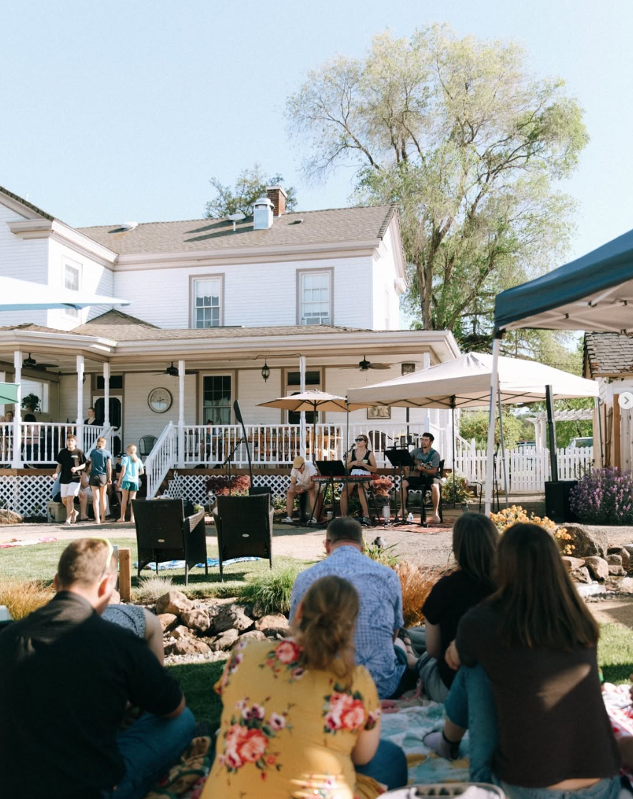 People gathered in a backyard, enjoying an outdoor event with musical performance on the patio of a large white house, under umbrellas, with trees and a clear blue sky.