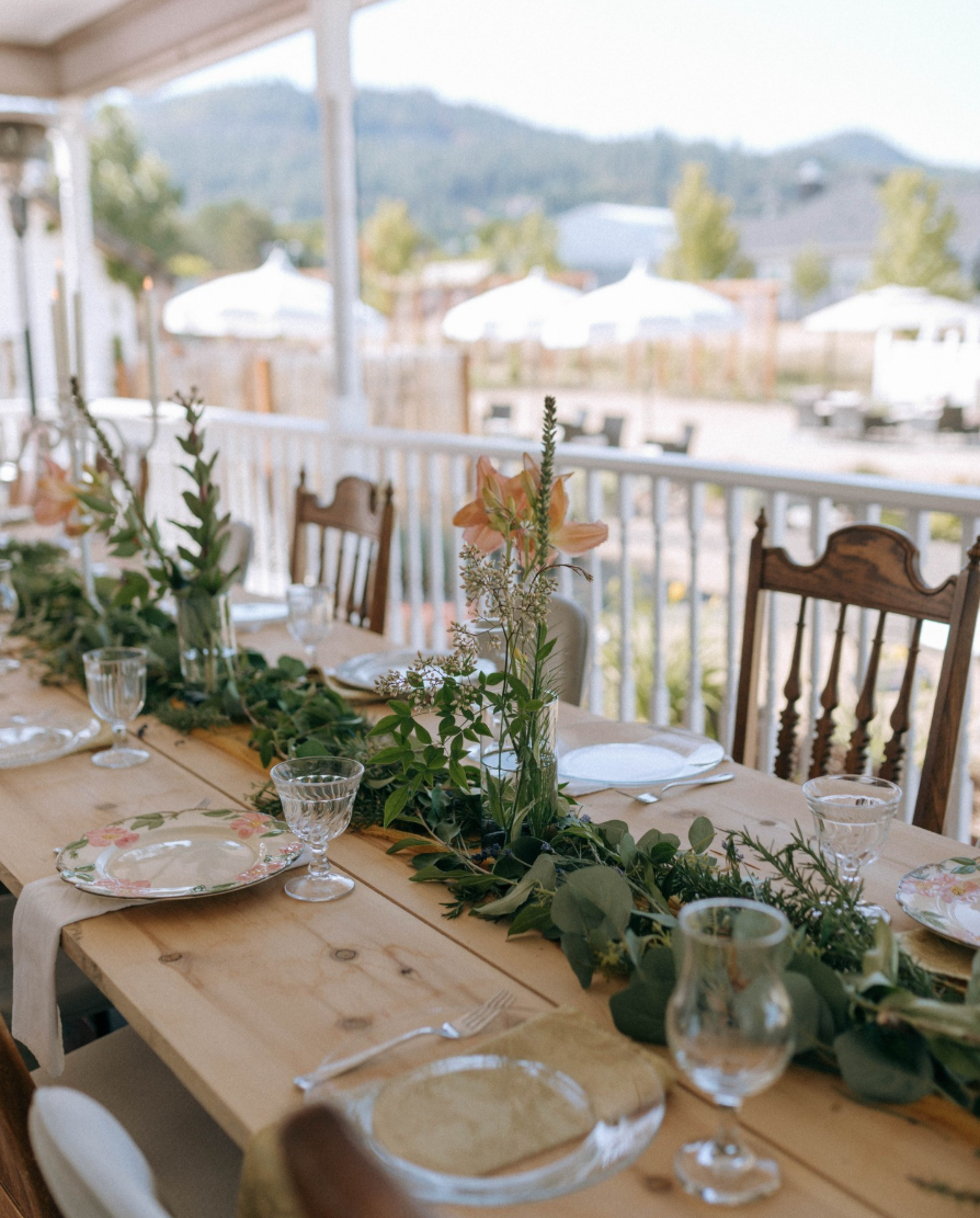 A wooden outdoor dining table set for a meal with floral plates, glassware, and cutlery, decorated with a green foliage and flower centerpiece, on a porch with a view of houses, umbrellas, and mountains in the background.