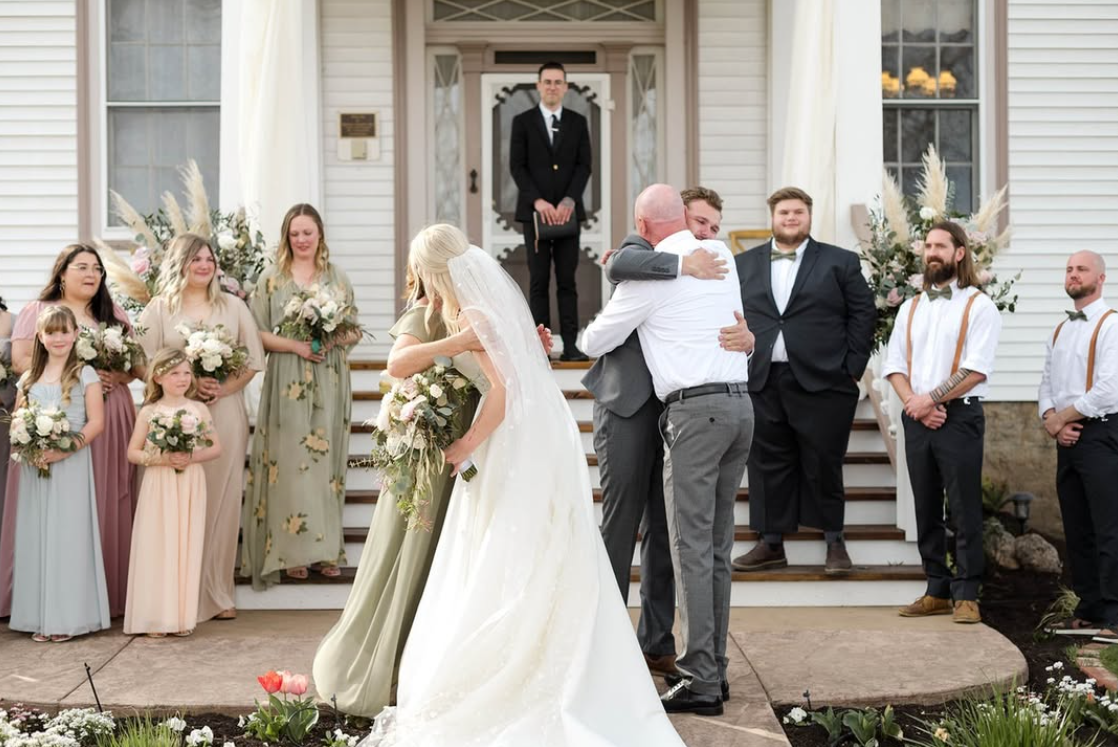 A wedding ceremony outside a church with the bride and groom hugging, surrounded by family and friends. Bridesmaids and groomsmen stand nearby, with the officiant in the doorway.