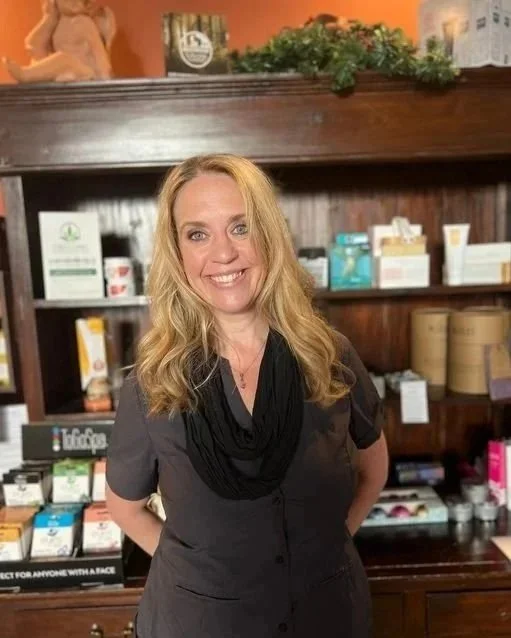 A woman with blonde hair, smiling, standing in front of a wooden display shelf with various health and wellness products in boxes and jars.
