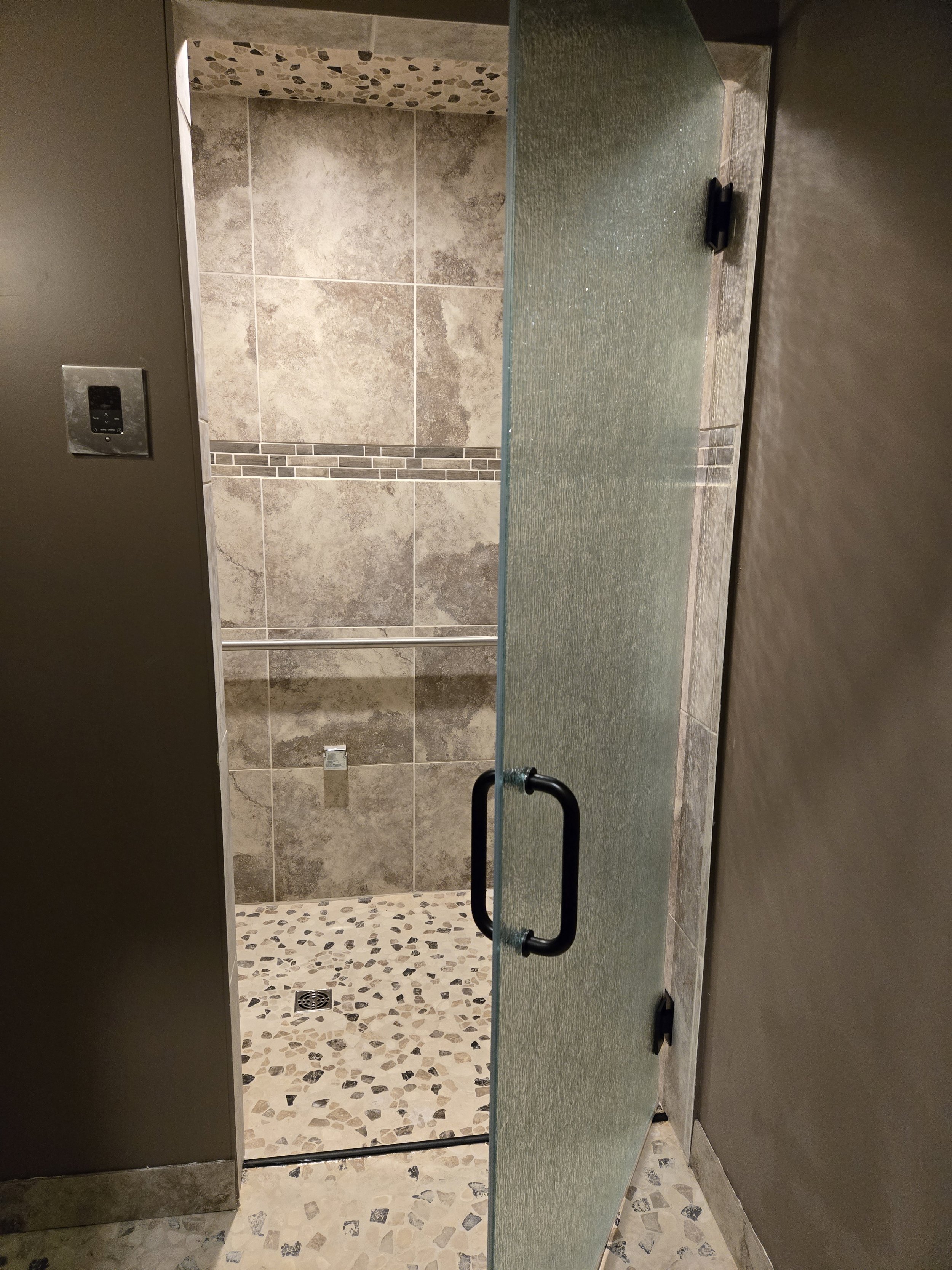 An open door leading into a women’s restroom stall with a frosted glass door and black handle, beige tiled walls with decorative mosaic tile accent, and a pebble-patterned floor with a drain visible at the bottom.