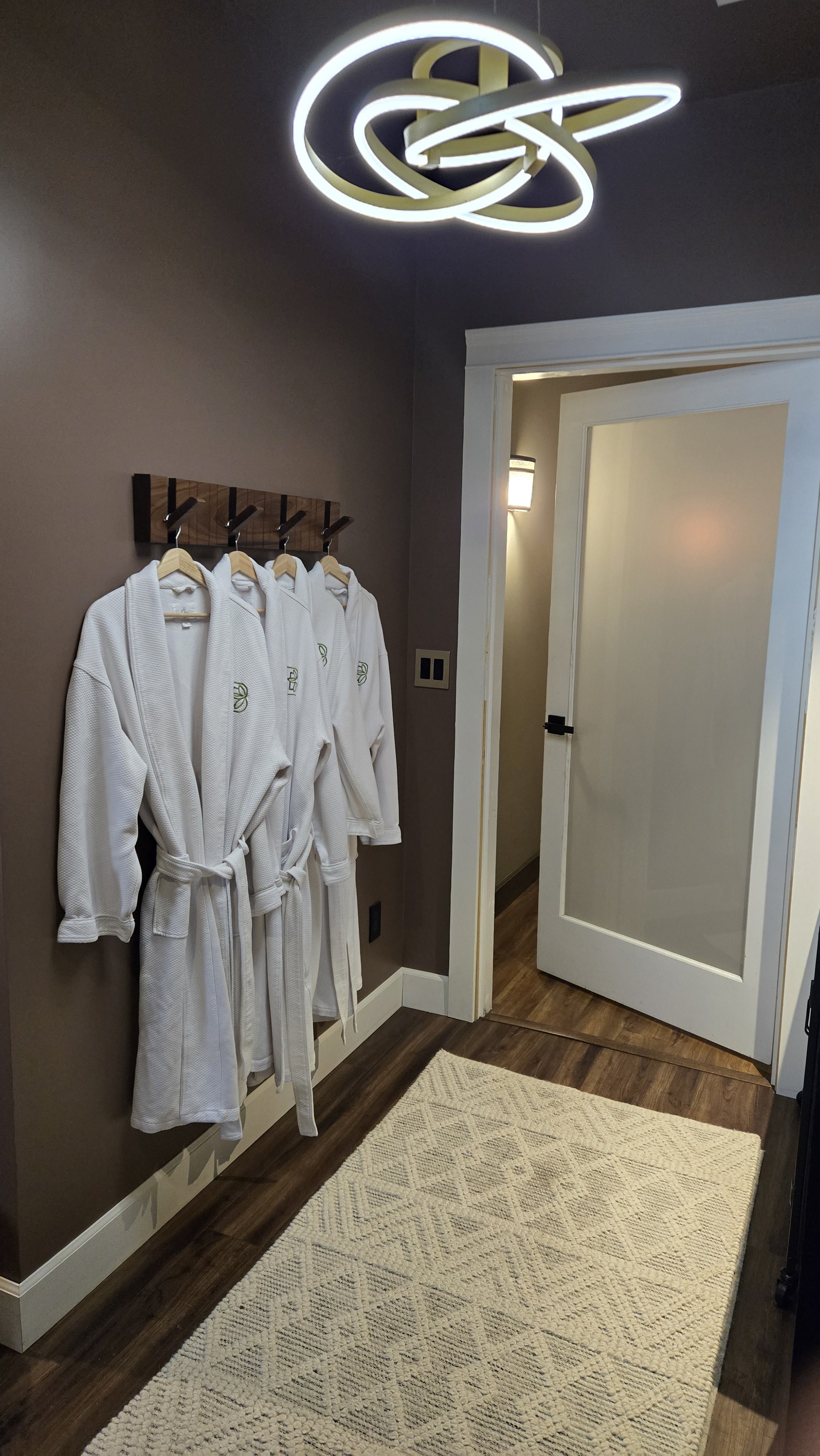 A hallway corner with four white bathrobes hanging on a wooden coat rack, a white door slightly open, a textured beige rug on dark wood flooring, and a modern ceiling light fixture with abstract loop designs.