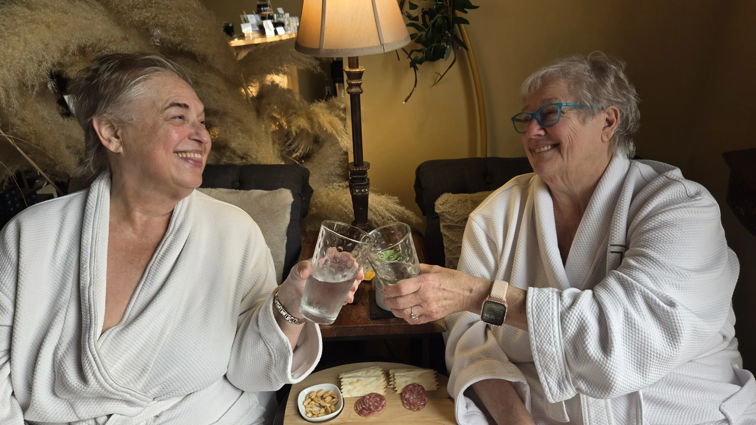 Two elderly women in white robes smiling and clinking glasses of water in a cozy living room. They are seated at a table with a charcuterie board of meat, cheese, and snacks, enjoying a friendly moment together.
