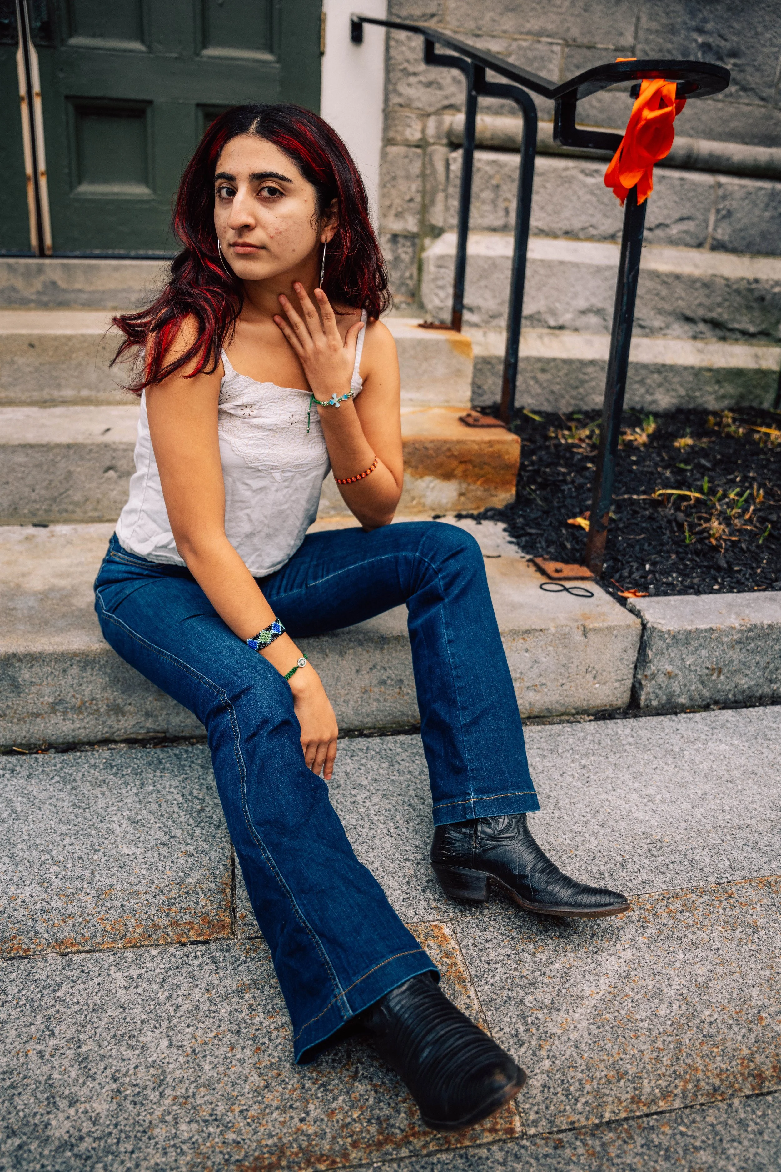 A young woman with dark hair and red highlights sitting on concrete steps outside a building, wearing a white sleeveless top, blue jeans, and black cowboy boots. She has jewelry on her wrists and a bracelet on her ankle, and is looking at the camera with a serious expression.