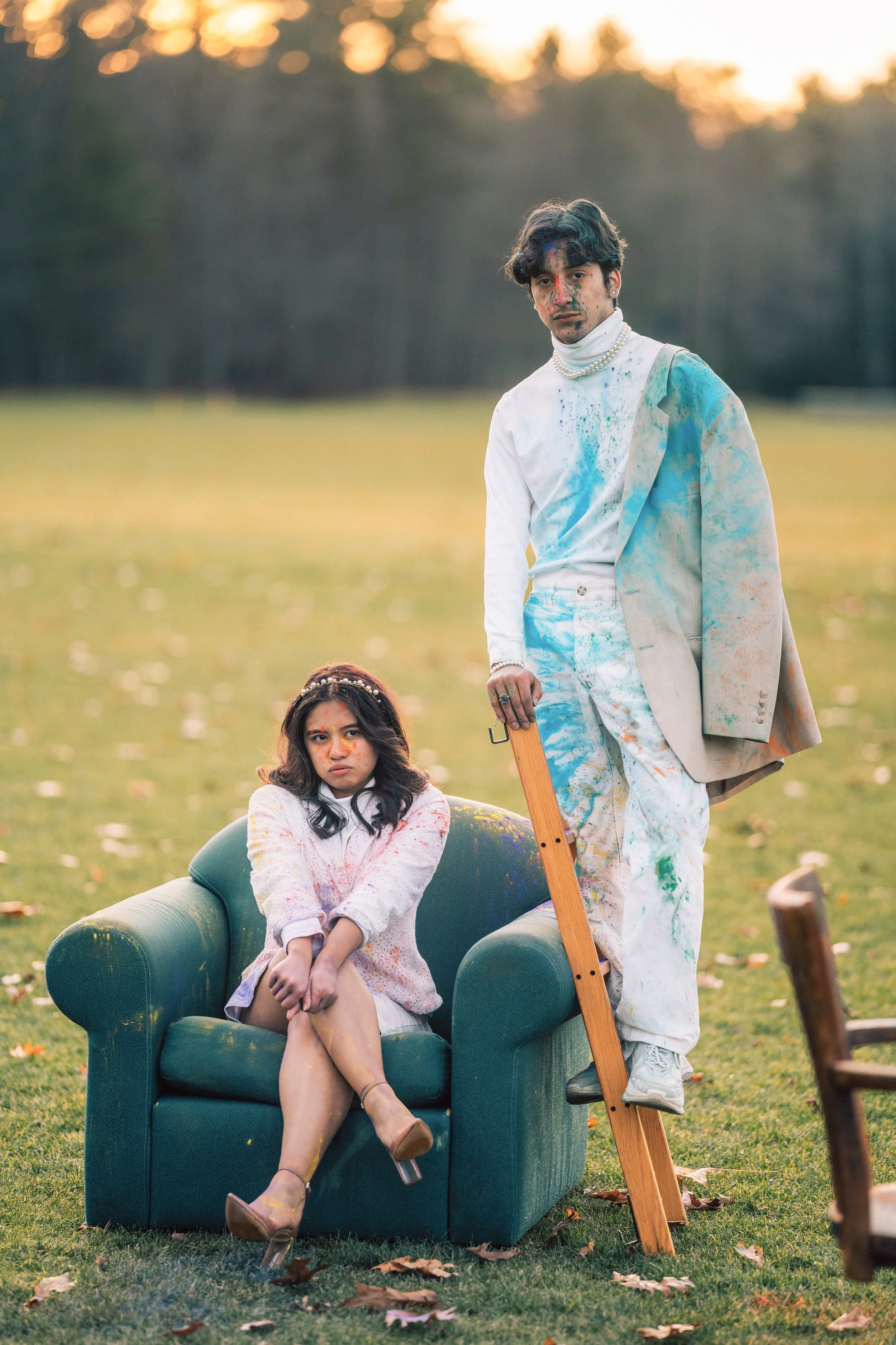 A young woman seated on a green sofa outdoors, with paint splatters on her face and clothes, and an older man standing next to her with a wooden ladder, both covered in colorful paint. The setting is a grassy field during sunset.