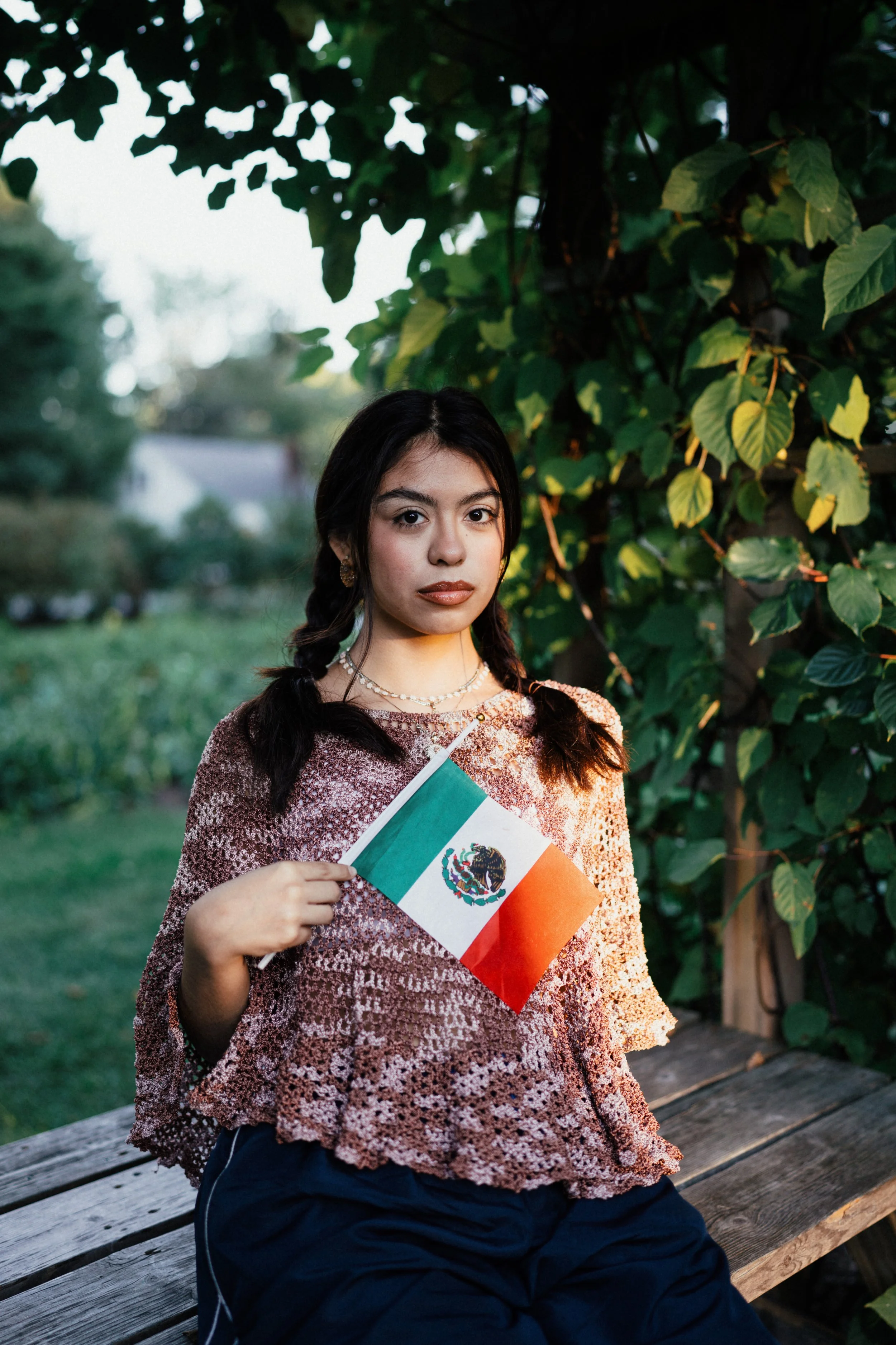 A young woman with dark hair in two braids, wearing a patterned top and dark pants, holding a small Mexican flag in front of a green leafy background, sitting on a wooden bench outdoors.