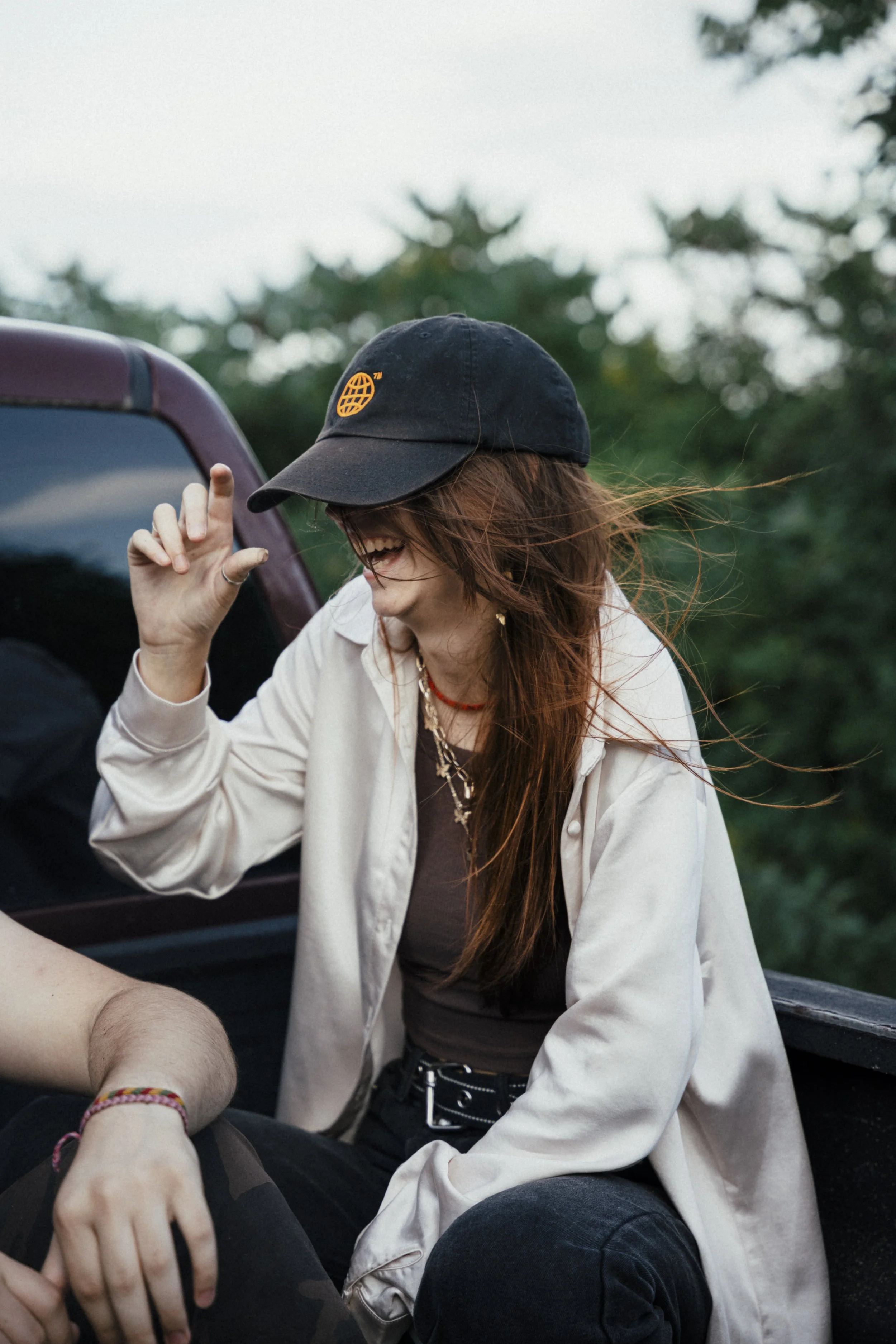 A woman with long red hair wearing a black cap and beige jacket, smiling and laughing outdoors near a vehicle.