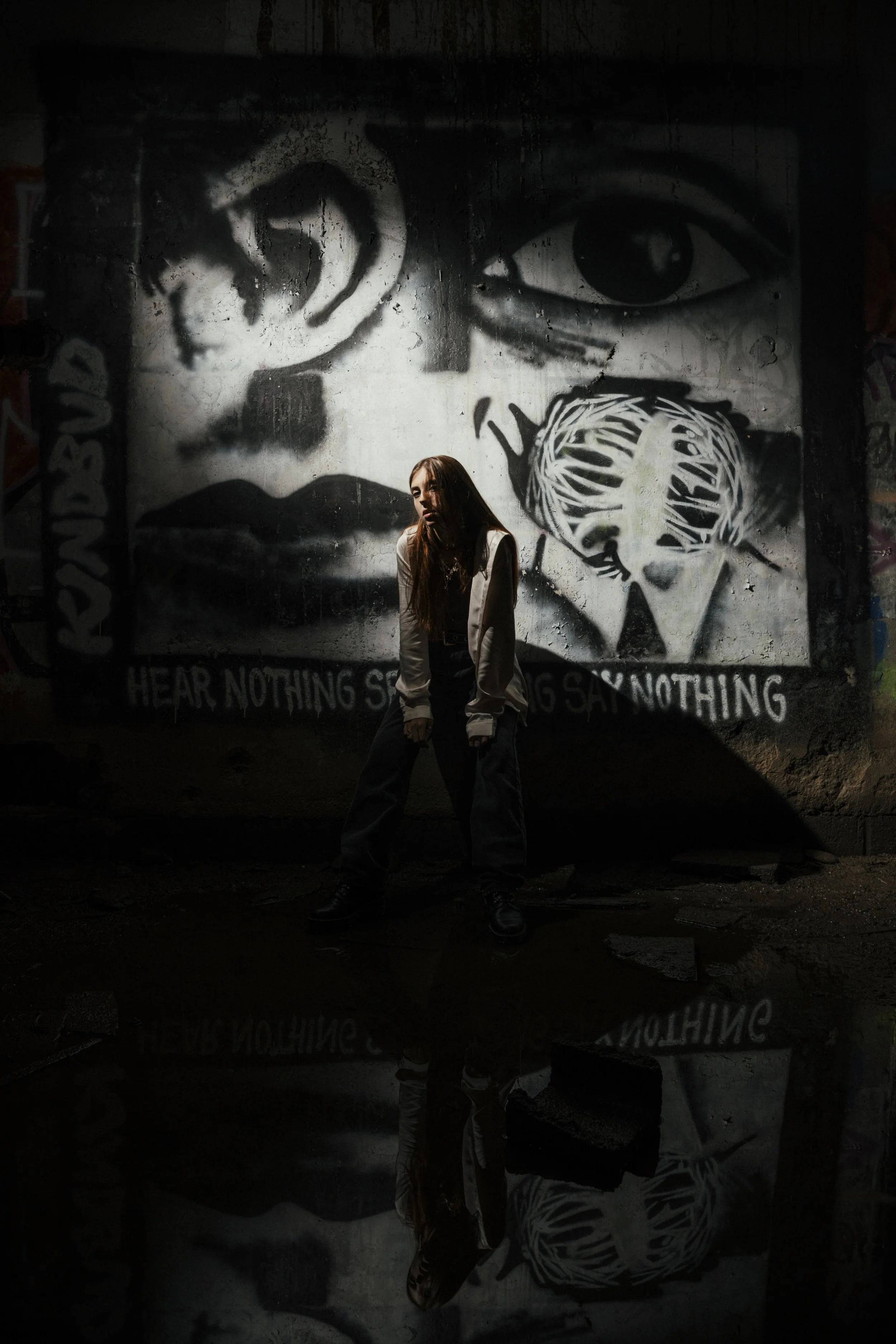 A young woman standing in front of a large black and white graffiti mural on a wall, which features a woman's face with one eye open and a stylized brain with lines crossing through it. There is text at the bottom of the mural that says "HEAR NOTHING SAY NOTHING". The woman is illuminated by a directed light, creating a shadow on the wall and a reflection on the wet floor.