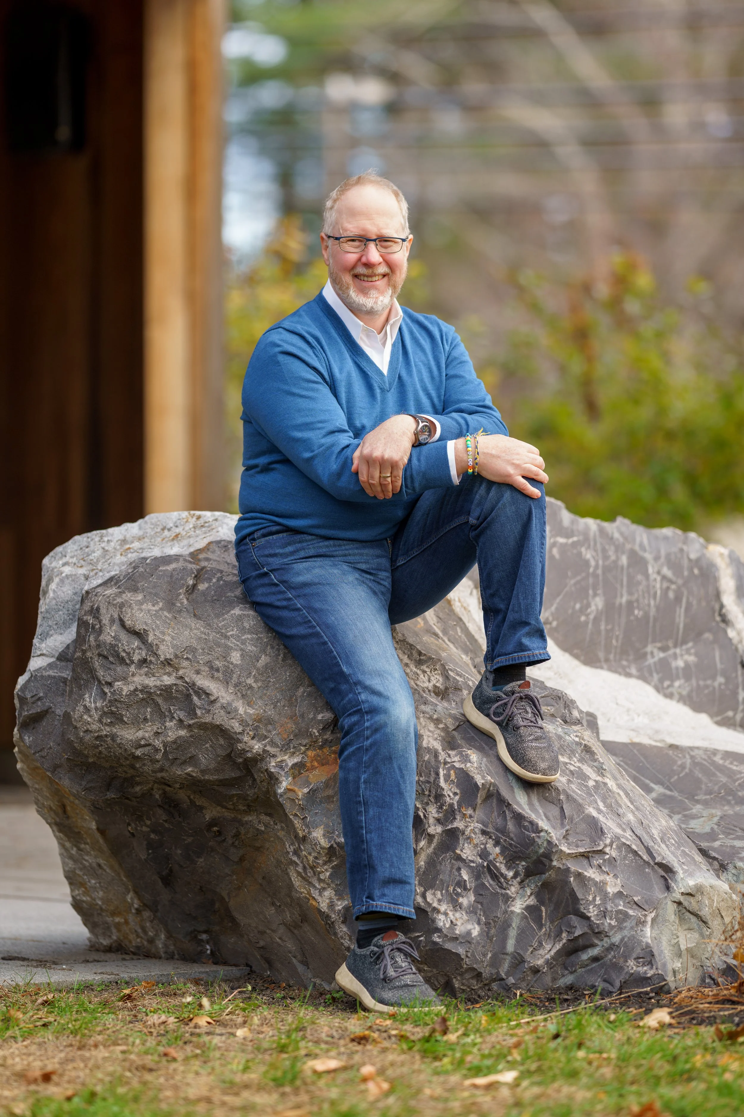 A man sitting on a large rock outdoors, smiling, wearing glasses, a blue sweater, white shirt, jeans, and sneakers.