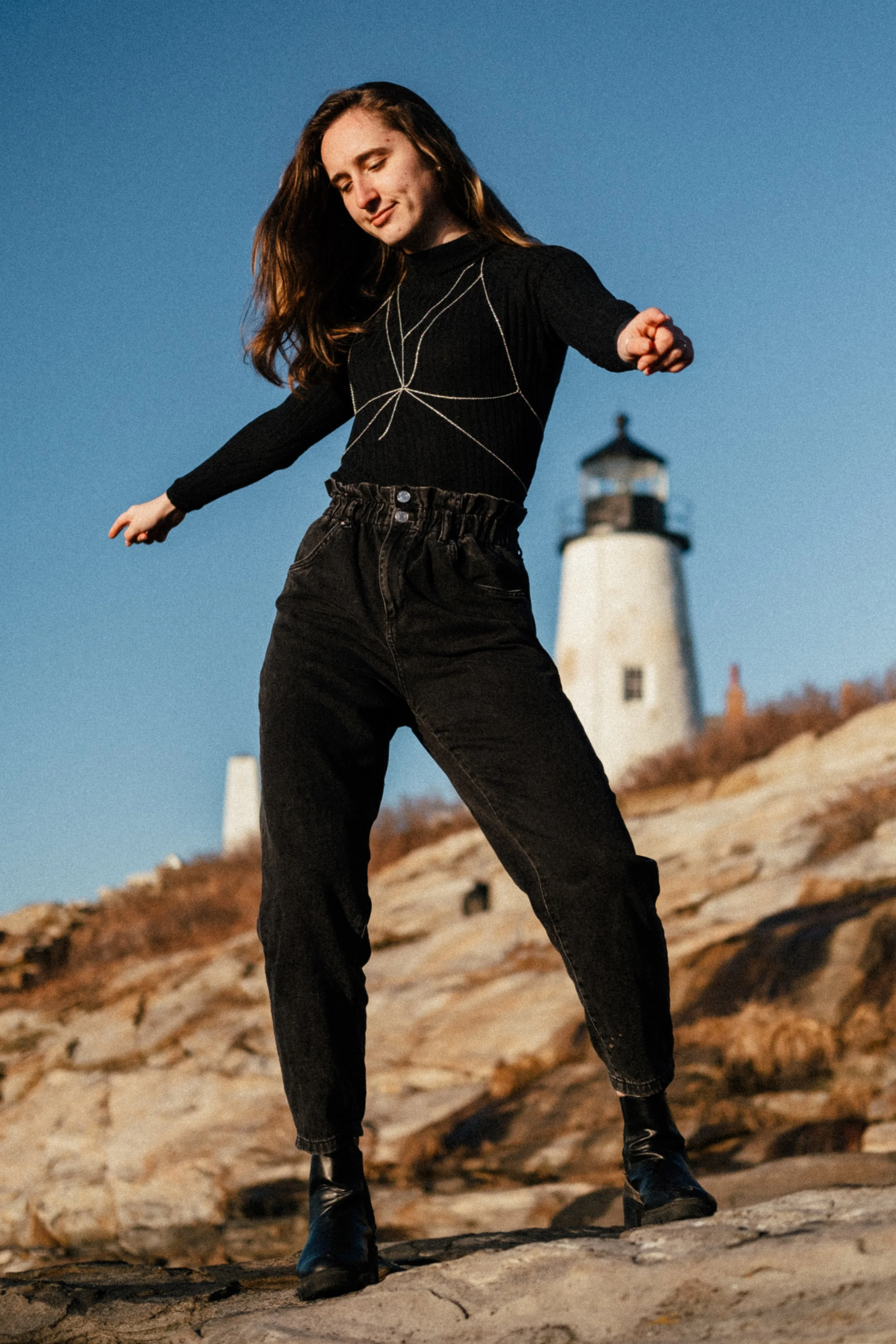 Woman standing on rocky ground with arms outstretched, tall lighthouse in background, clear blue sky.